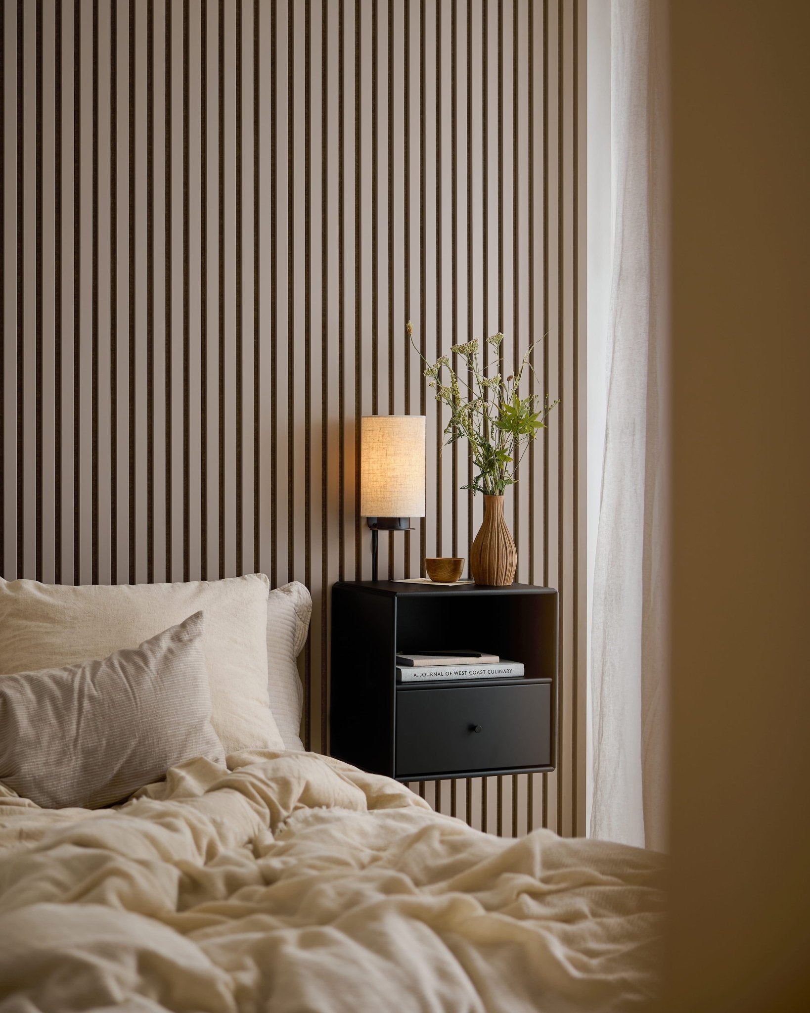 Uma cama com roupa de cama creme ao lado de uma mesa de cabeceira preta, em frente a uma parede de Akupanel em Dark Cashmere.