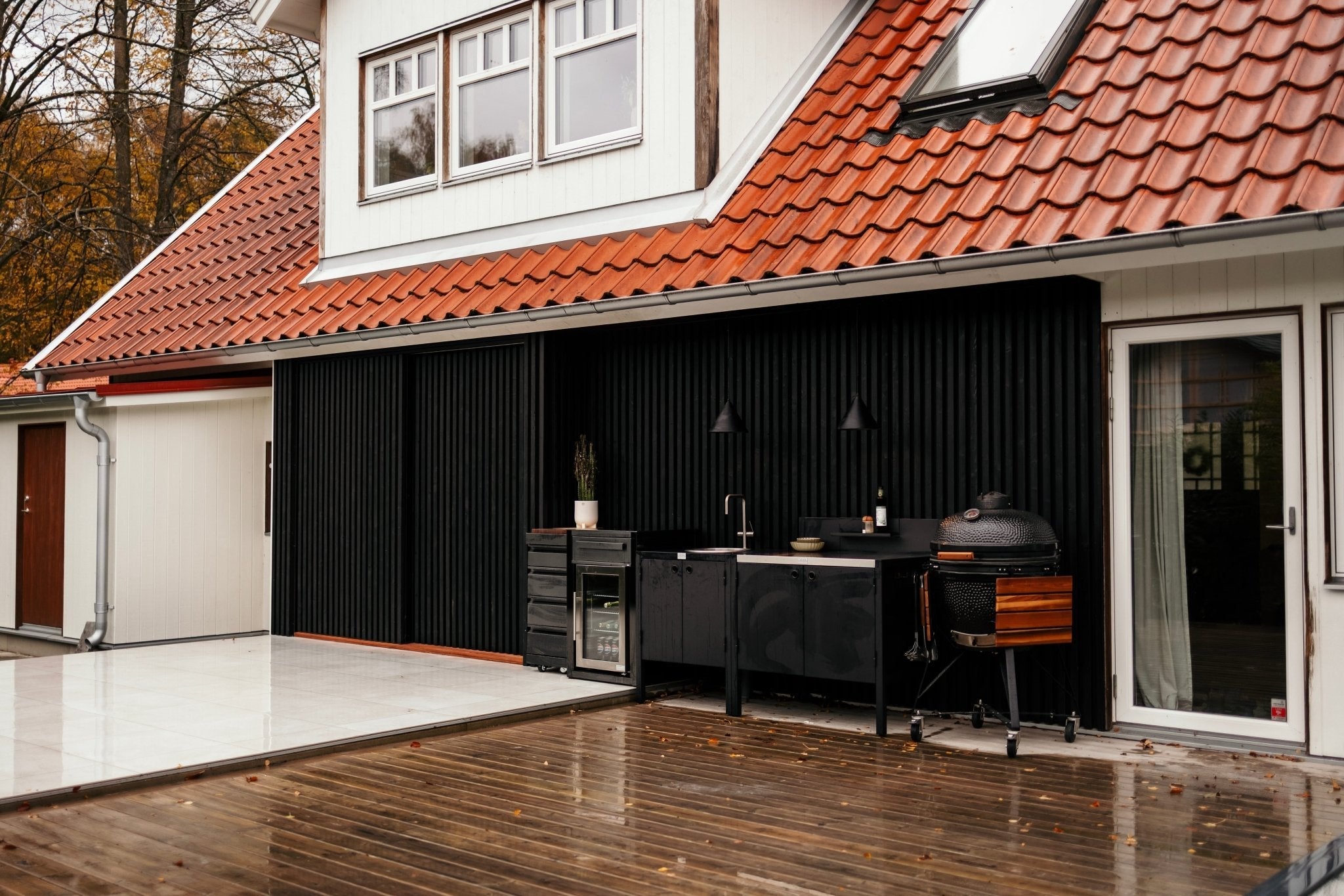 Black slatted wall under a red tile roof, closed, behind an outdoor kitchen with a sink, two pendant lamps and a black grill on a wet wooden deck