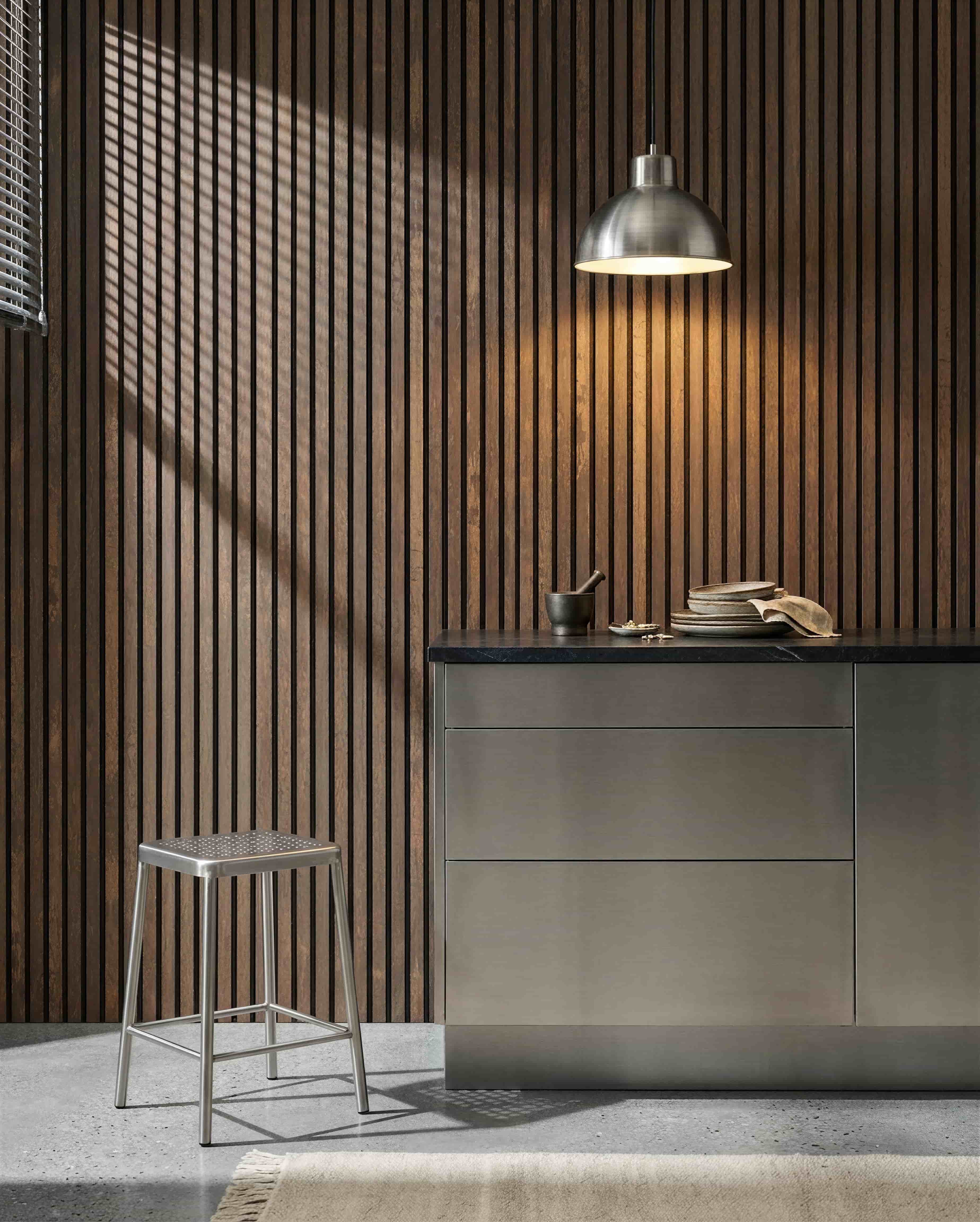 Cozinha com uma parede acústica ripada WoodUpp Akupanel Oxide Copper, bancada em aço inoxidável, banco metálico, candeeiro suspenso, louça variada e almofariz com pilão.