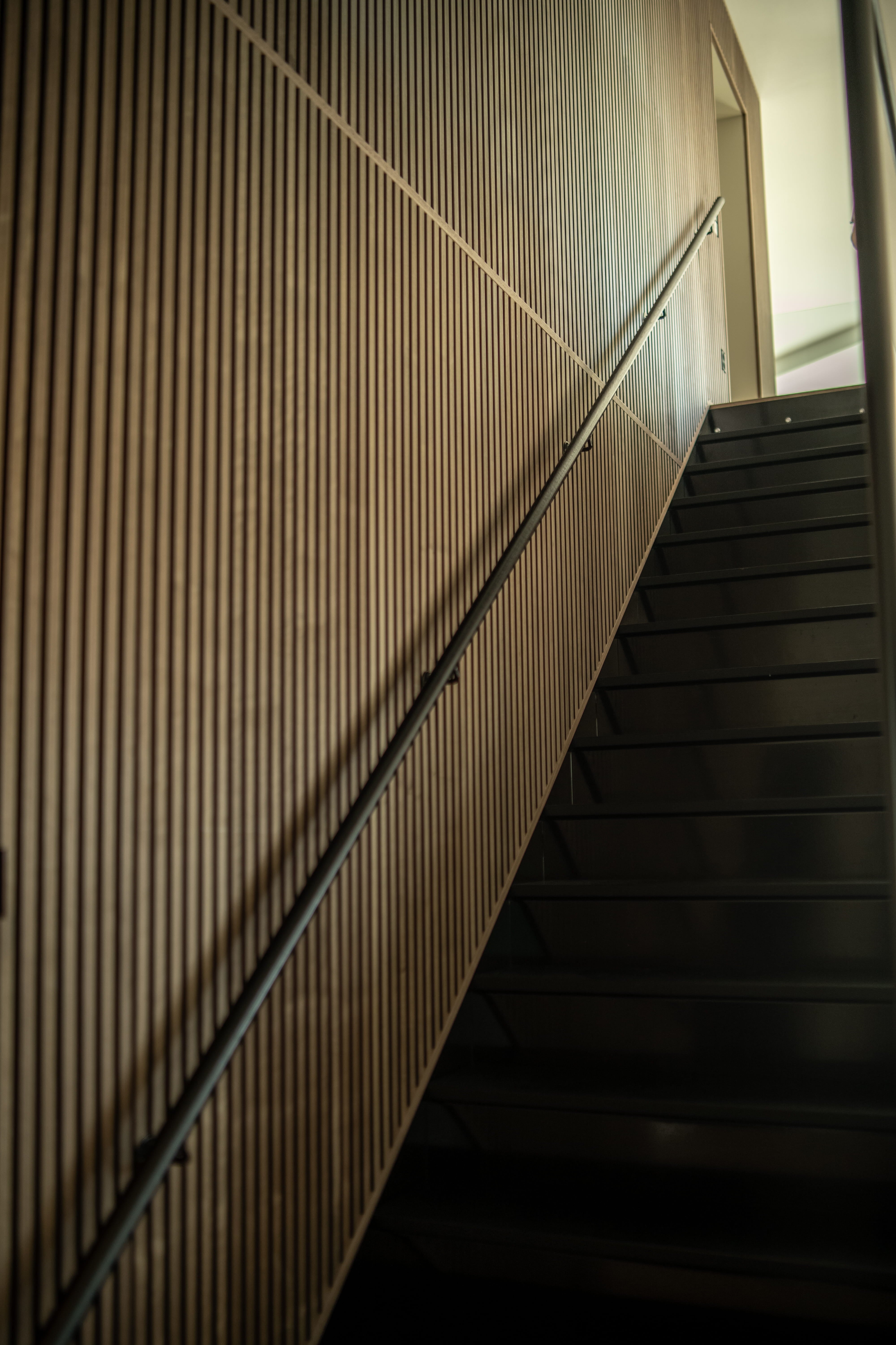 Uma escada moderna com degraus pretos é complementada por um painel acústico vertical de parede de ripas, visto de baixo em um ângulo para uma aparência nórdica minimalista.