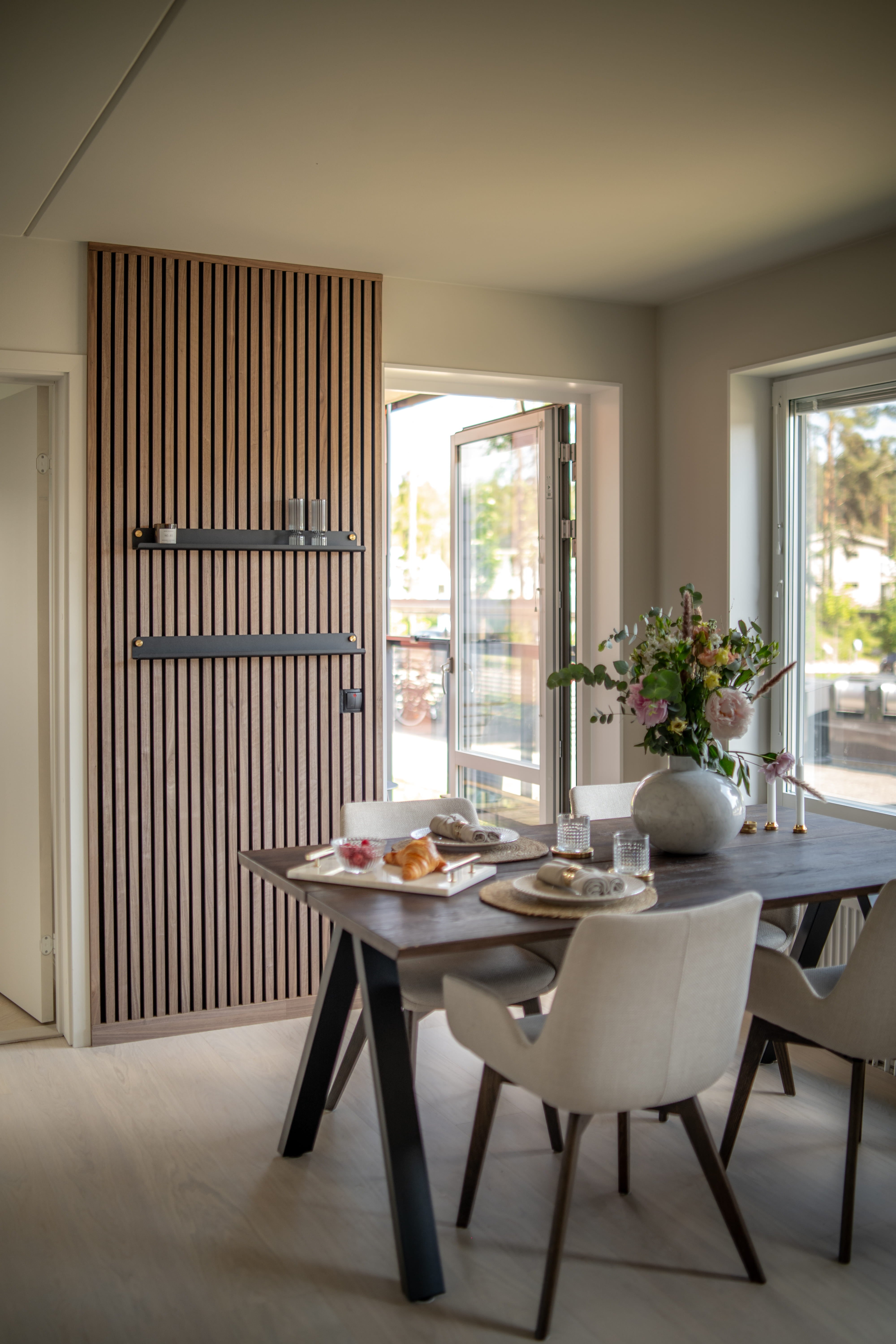 Sala de jantar moderna com parede de ripas de madeira acústica Akupanel texturizada, mesa de café da manhã escura, cadeiras bege minimalistas e um vaso de flores ao lado de grandes janelas abertas.