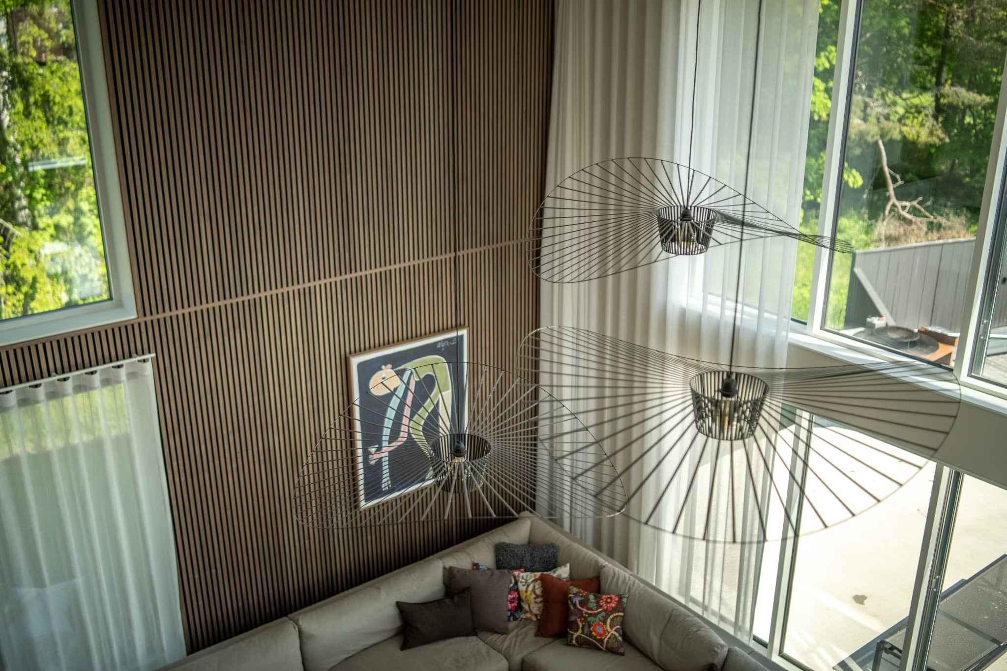Sala de estar moderna com paredes de madeira nórdica Akupanel, janelas amplas, arte abstrata em painel acústico, um sofá seccional e luminárias minimalistas com estrutura de arame, vista de cima.