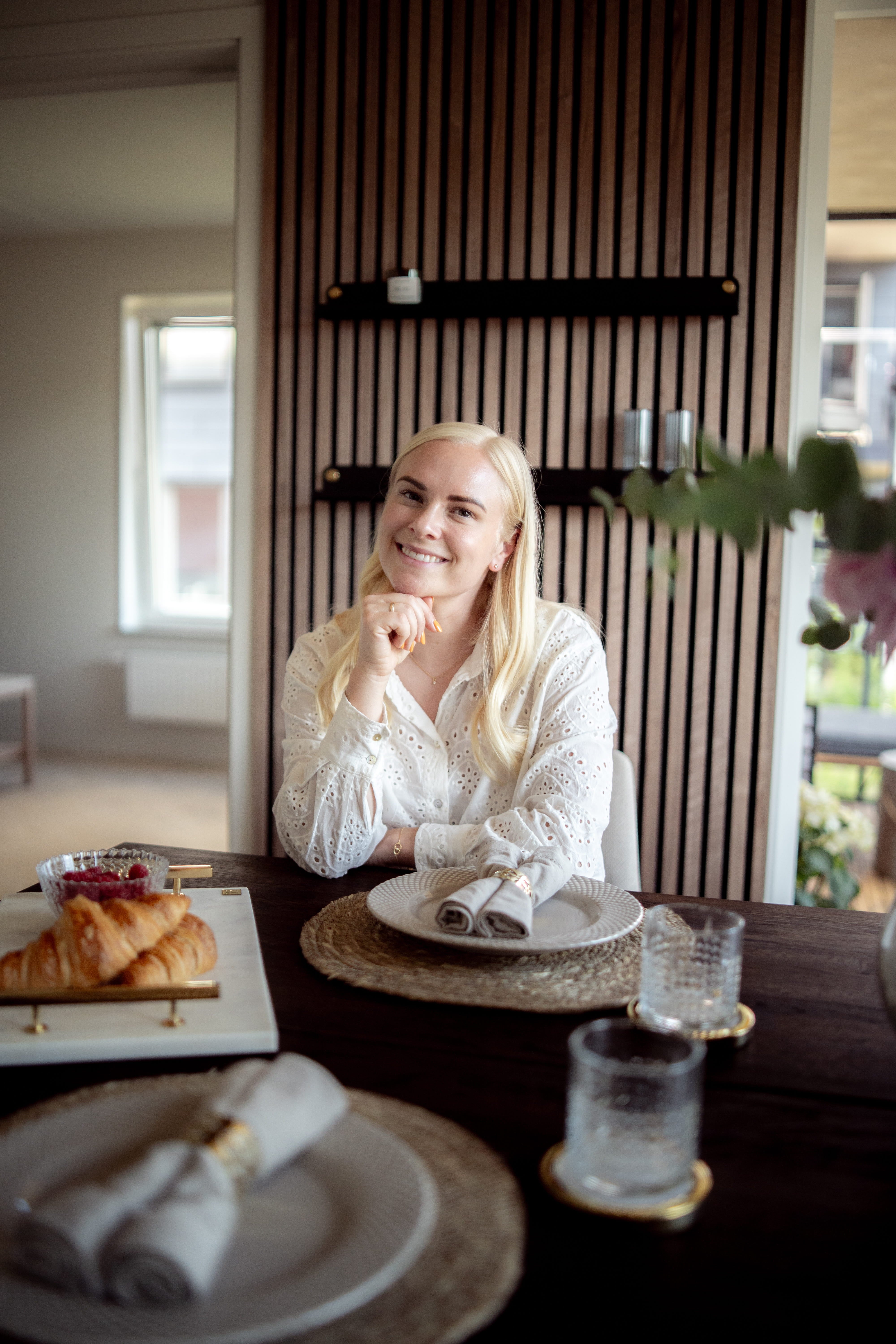 Uma mulher com longos cabelos loiros está sentada em uma mesa de jantar preparada para o café da manhã, com croissants e copos, ao lado de um painel acústico nórdico na parede.