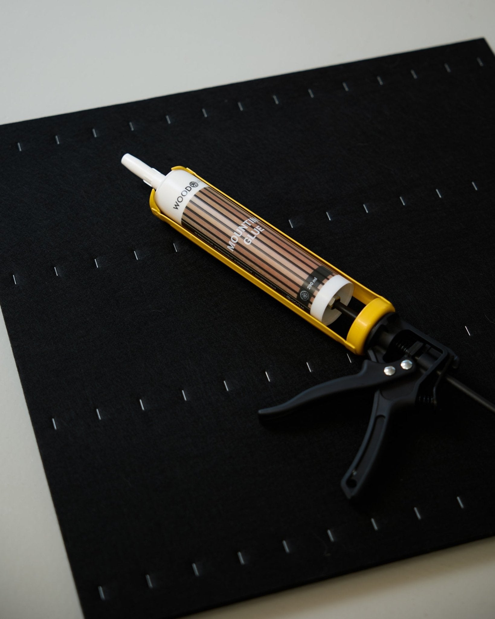 Uma WoodUpp Grout Gun amarela e black (290 ml) está pousada sobre um tapete black com uma grelha de agrafos brancos, usada na instalação de painéis acústicos.