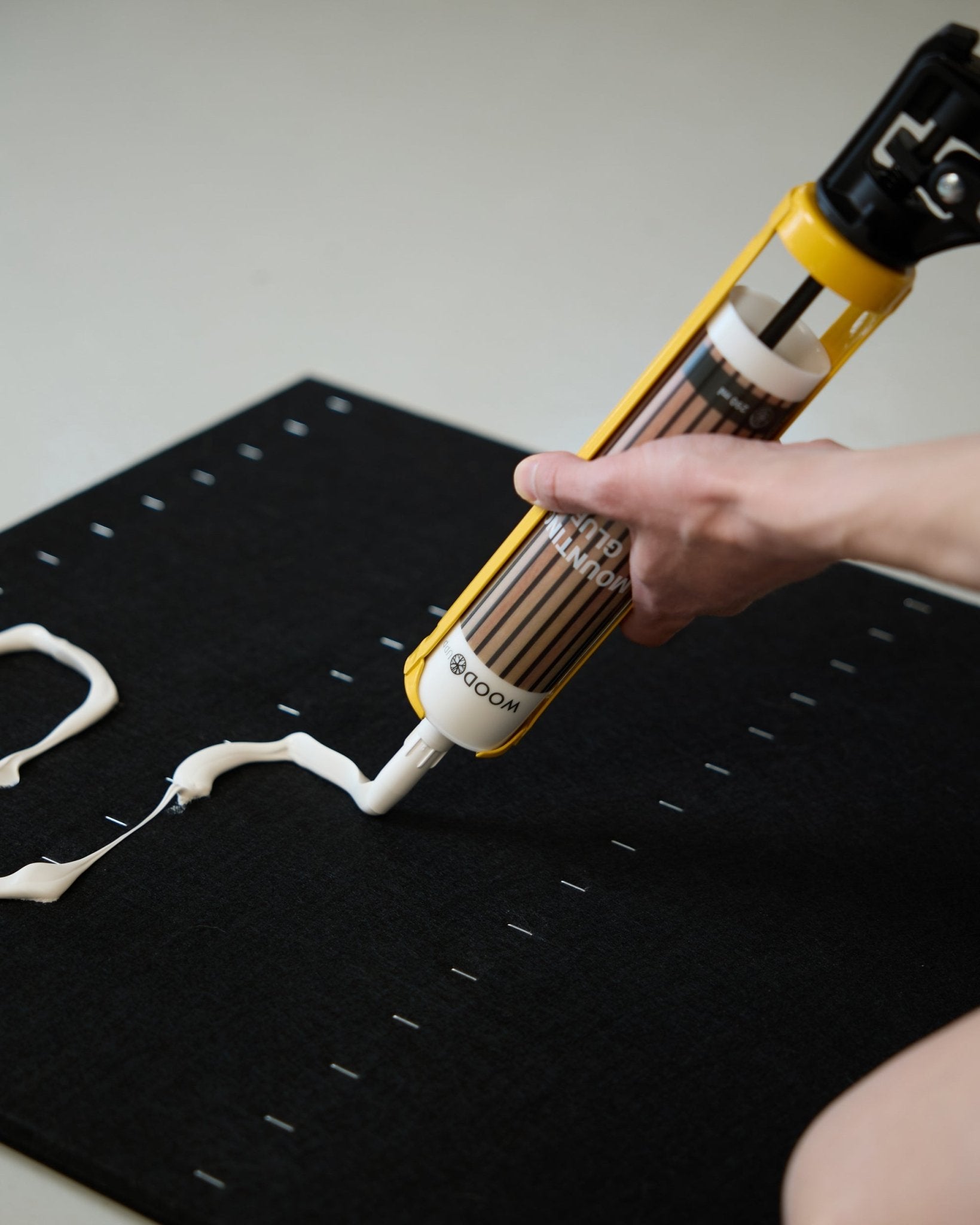 Uma mão aplica cola de montagem branca em padrão zig-zag com a WoodUpp Grout Gun (290 ml) sobre uma superfície black Painel acústico.