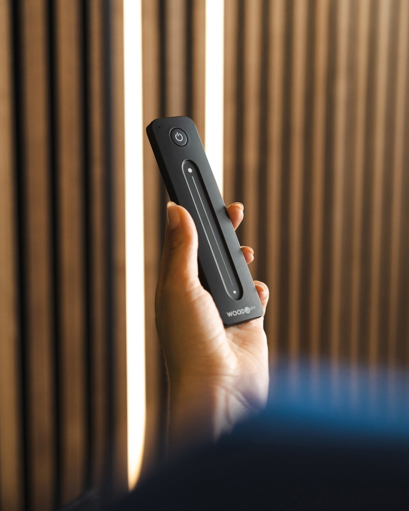 Uma mão segura um comando para fita LED black WoodUpp Lumi com botão de ligar/desligar, colocado em frente a painéis acústicos ripados WoodUpp; compatível com Philips Hue.