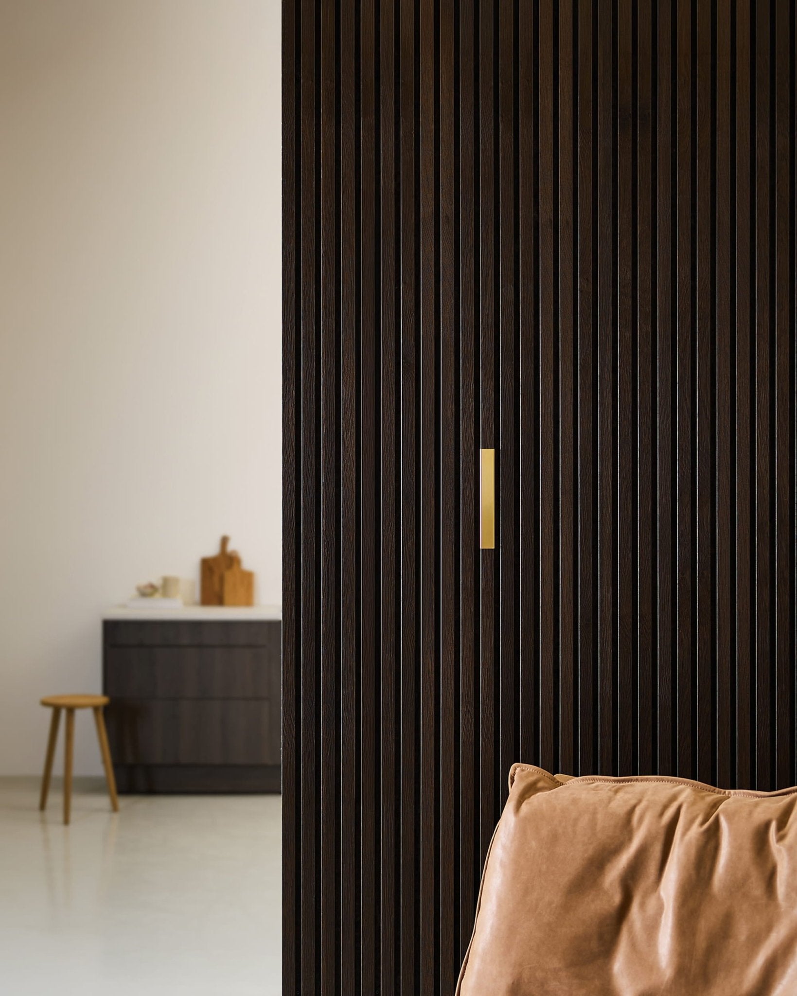 Quarto minimalista com um acessório WoodUpp Liva | Cover / Latão, de 20 cm, numa divisória Akupanel de madeira escura, uma cadeira de couro castanho e um armário preto.