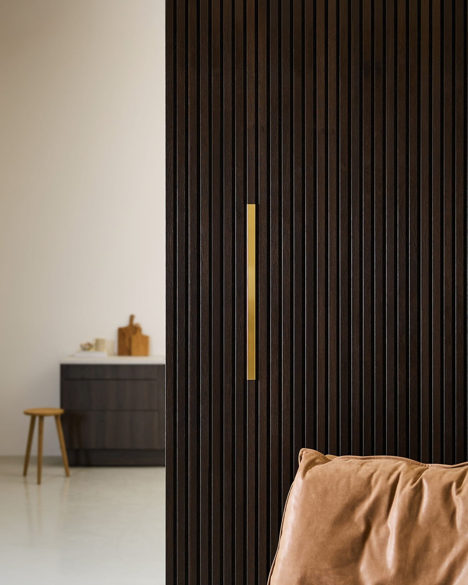 Quarto minimalista com um WoodUpp Akupanel Liva | Revestimento / Latão, 60 cm, ao lado de uma cadeira de couro, um banco e um armário escuro, num ambiente neutro.