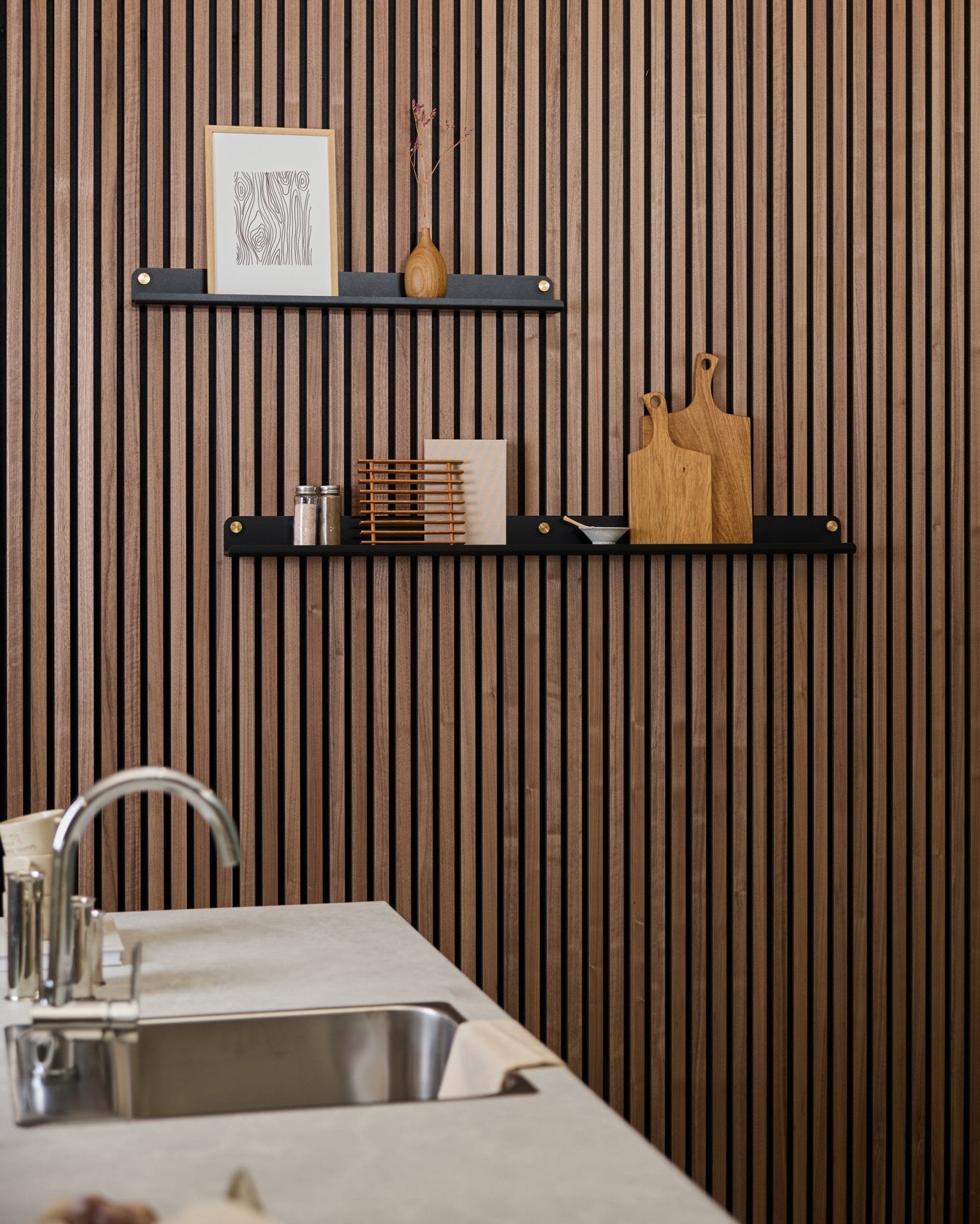 Cozinha minimalista com uma parede acústica ripada em madeira vertical Akupanel, prateleiras black, tábuas de corte, peças decorativas small e uma bancada com lava-loiça moderna à frente.