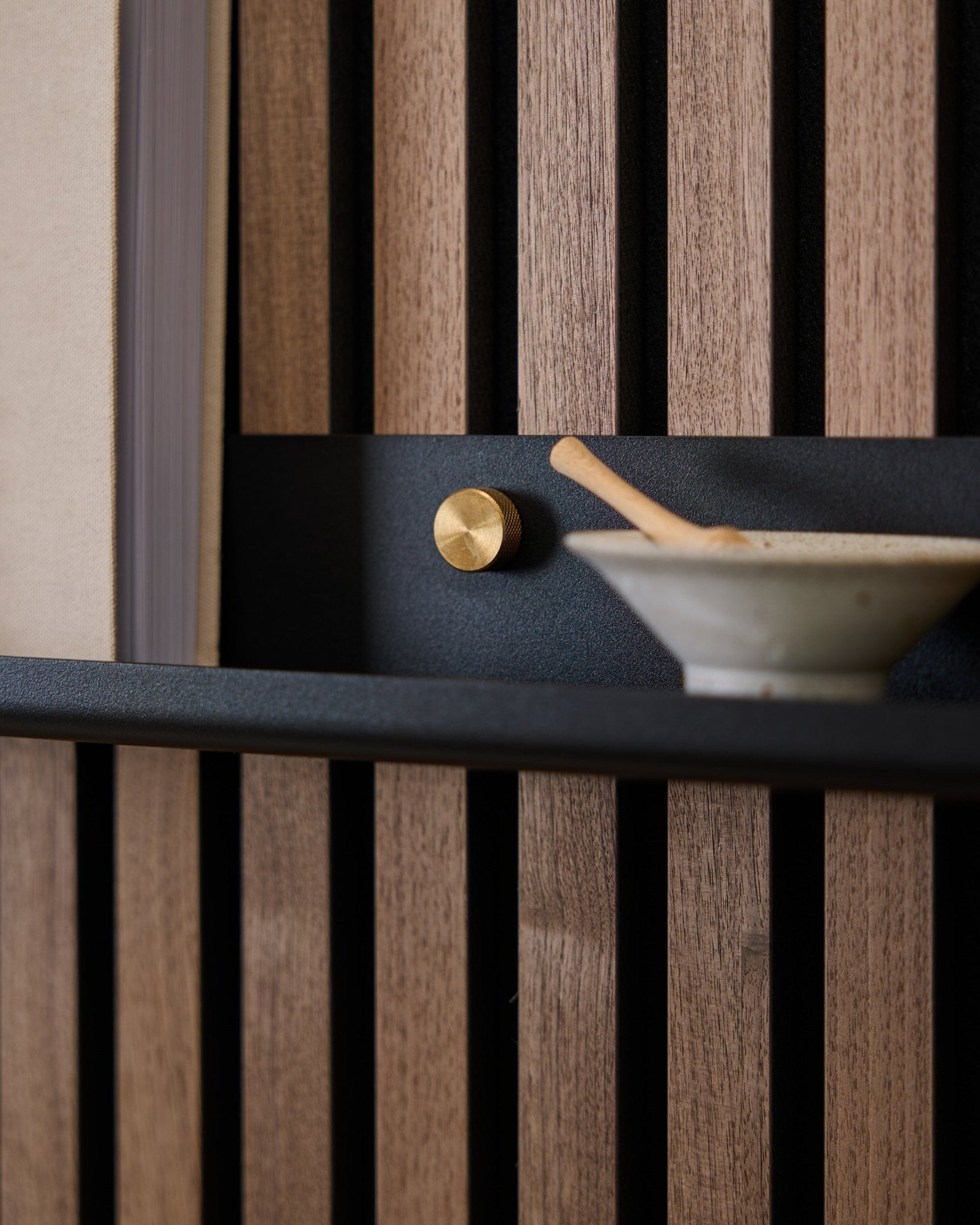 Grande plano de uma Narrow Shelf black WoodUpp Norr de 115,9 cm montada num painel acústico ripado nórdico com parafusos de aperto manual brass, botão dourado e taça de cerâmica com colher.