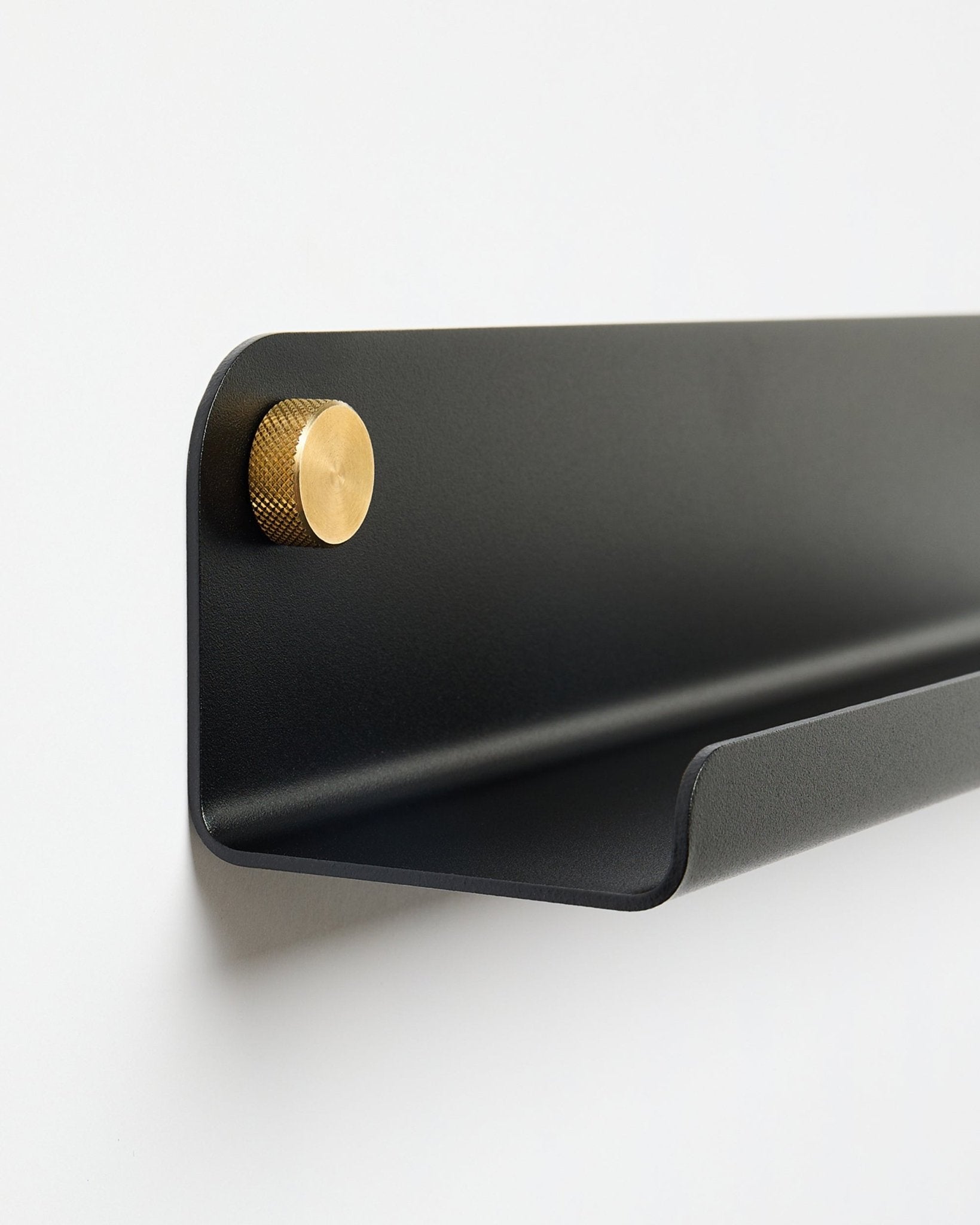 A Norr | Narrow Shelf em black (115,9 cm), com um único botão dourado à esquerda, da WoodUpp, montada numa parede branca lisa.