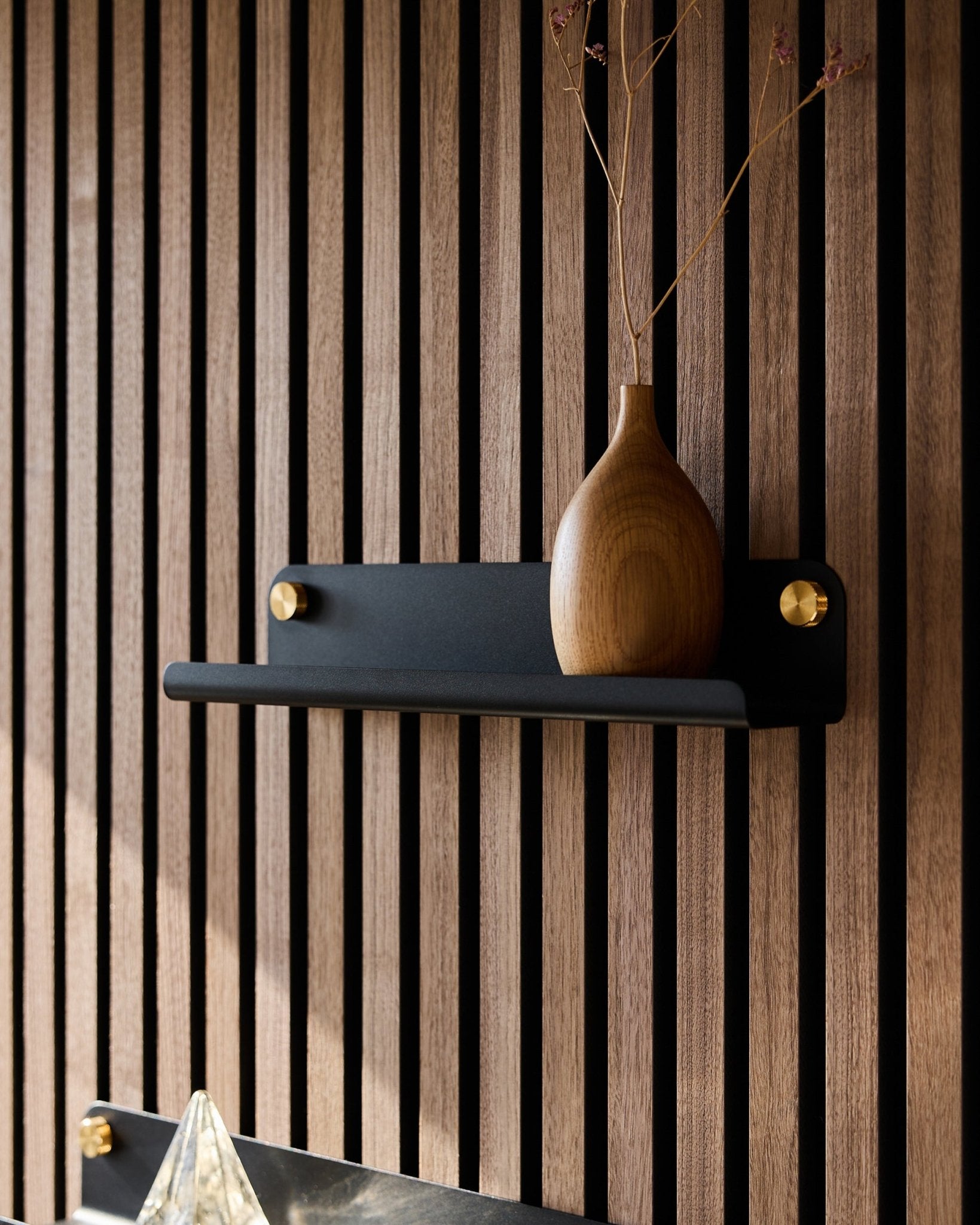 Uma jarra de madeira com ramos secos sobre uma prateleira estreita black WoodUpp Norr (35,9 cm), colocada diante de um painel acústico ripado em estilo vertical.