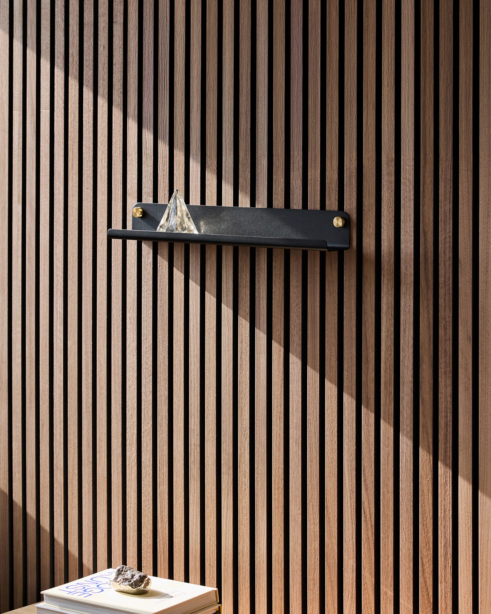Uma prateleira estreita Norr preta da WoodUpp, assente num painel acústico de madeira Akupanel, suporta uma pirâmide de vidro; a luz do sol incide sobre os livros que se encontram por baixo, encimados por uma pedra.