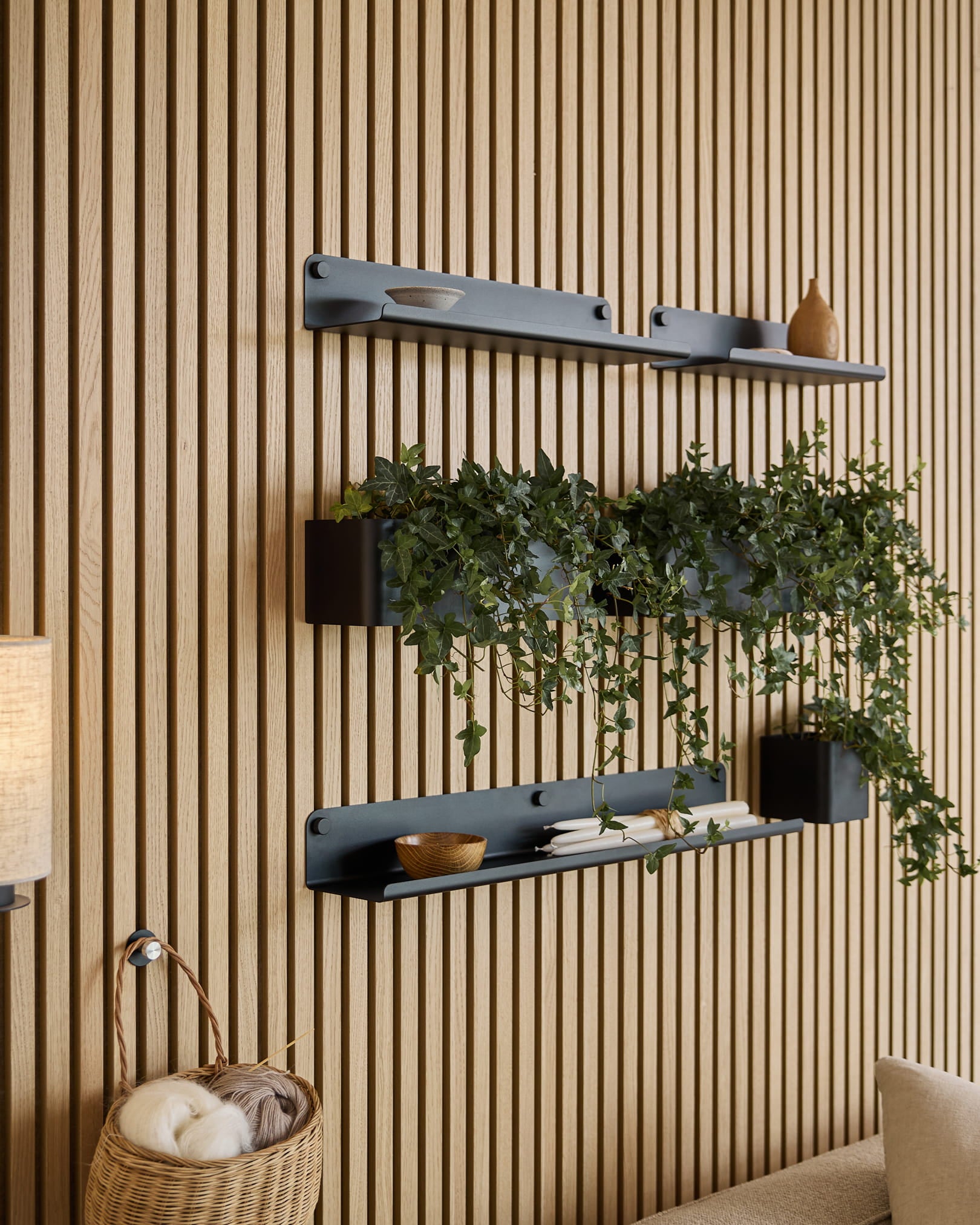 Três prateleiras black Norr | Plant Box da WoodUpp, com decoração e uma planta, estão montadas por cima de um cesto de verga com novelos de lã, sobre um painel acústico ripado verde.