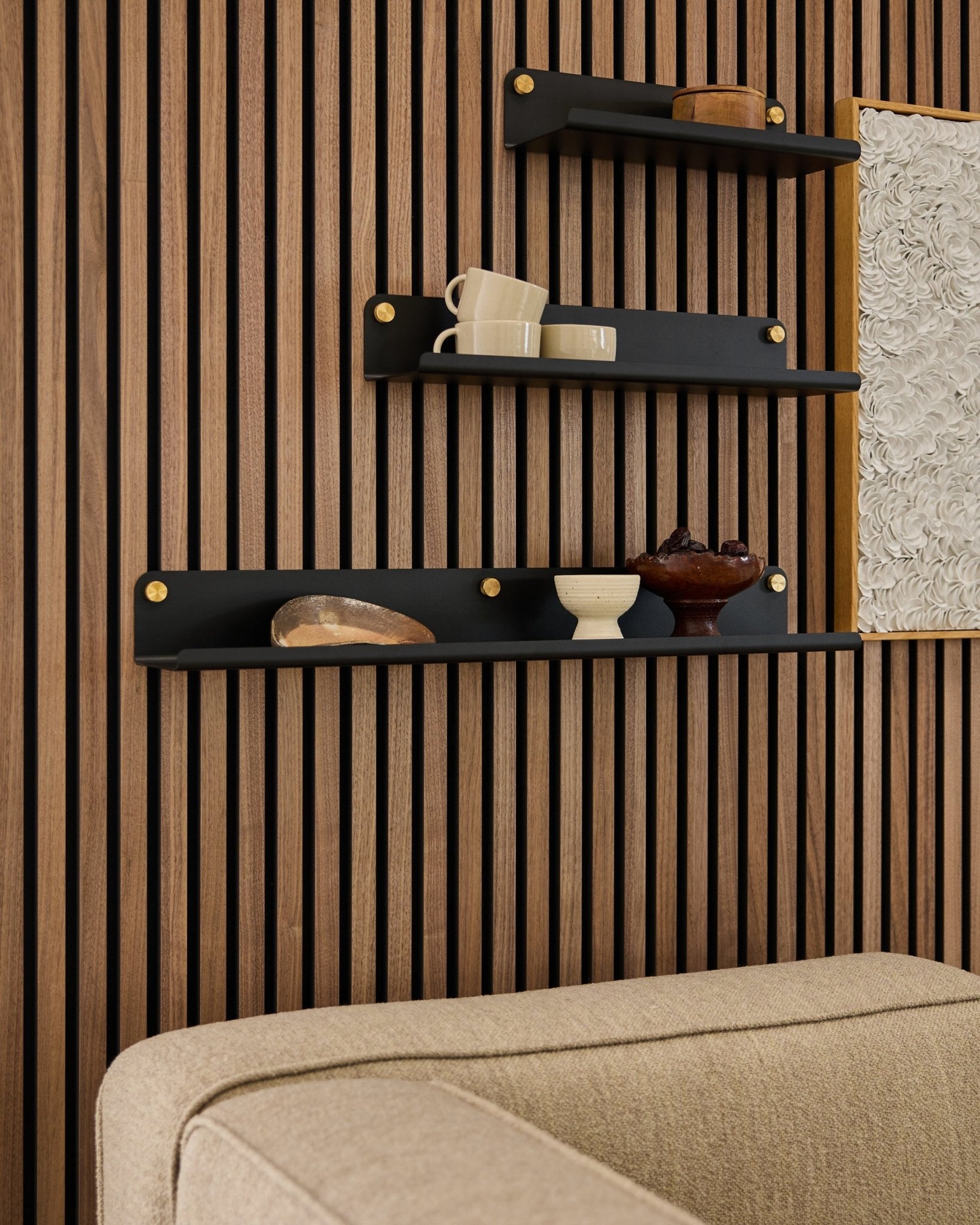 Três prateleiras black, incluindo a prateleira larga WoodUpp Norr (75,9 cm), exibem objetos decorativos numa parede Painel acústico de ripas de madeira, por cima de um sofá beige; parte da obra de arte está visível.