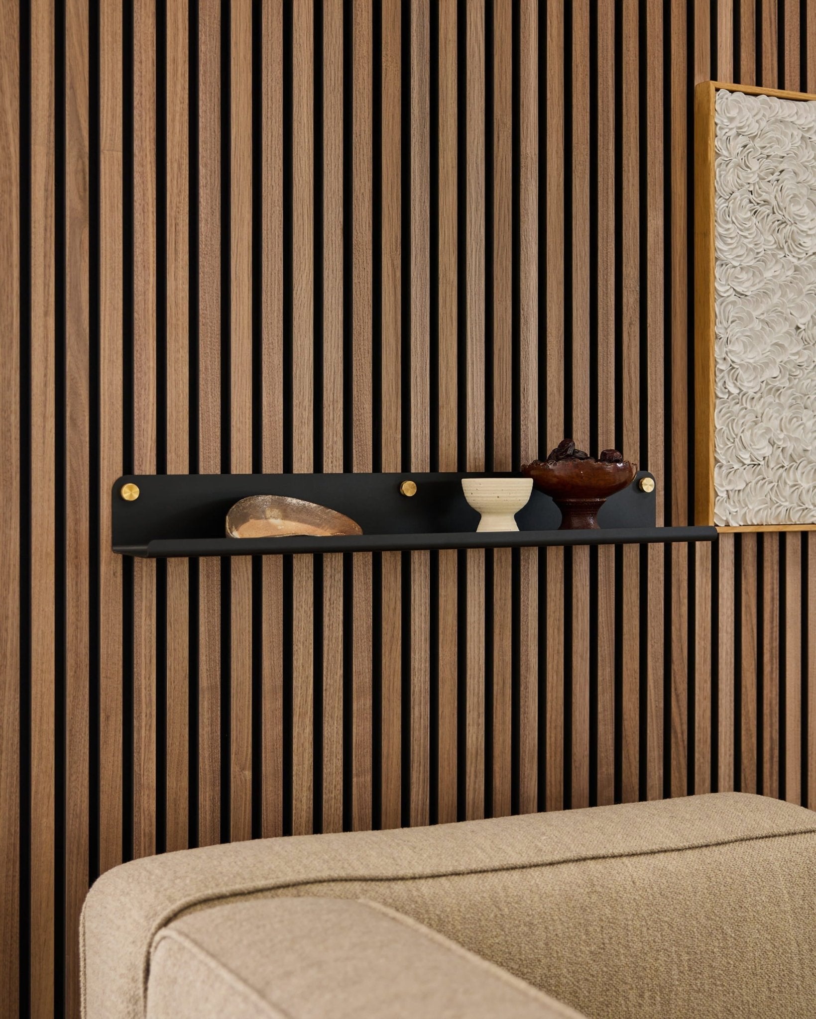 Prateleira larga WoodUpp Norr preta (75,9 cm) com decoração por cima de um sofá bege, fixada num painel de parede moderno de ripas acústicas em madeira.