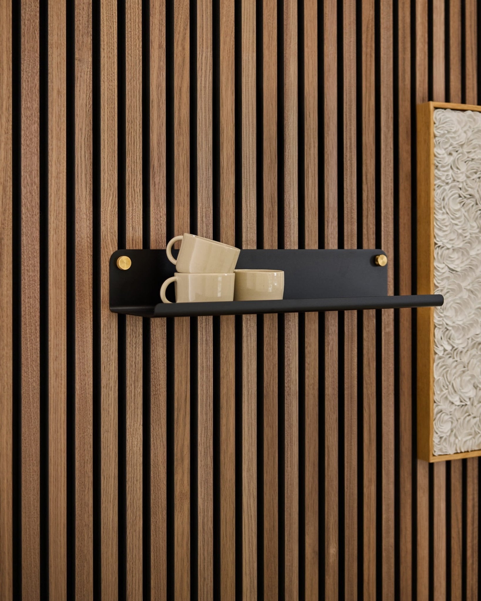 Uma prateleira Norr Wide preta da WoodUpp suporta três canecas bege, fixadas num painel acústico Akupanel ao lado de uma obra de arte branca texturada emoldurada.