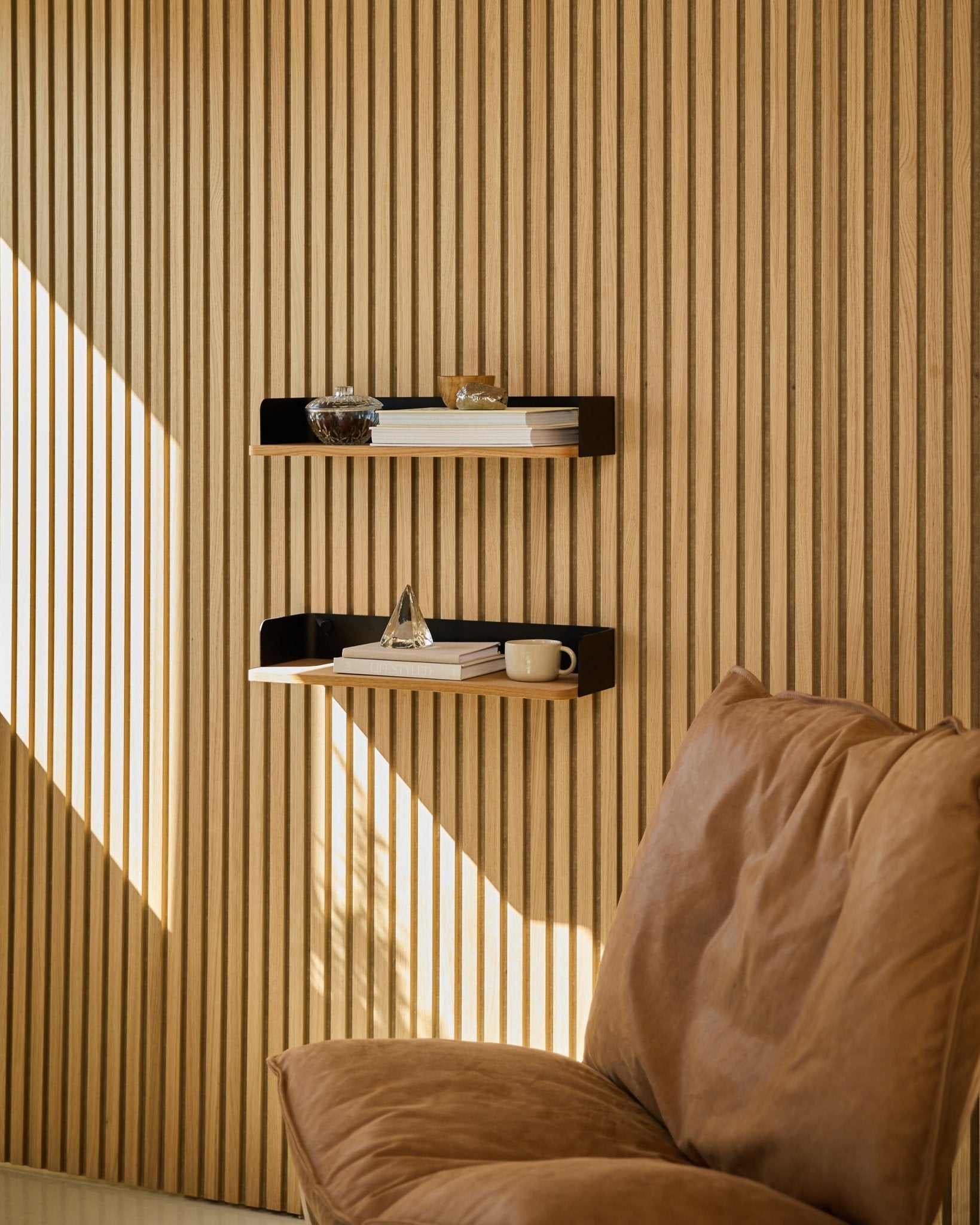 Cadeira Brown ao lado da estante Linn Bookshelf da WoodUpp em Natural Oak, com livros e decoração dispostos à luz natural junto a uma parede de ripas de madeira nórdica Painel acústico.