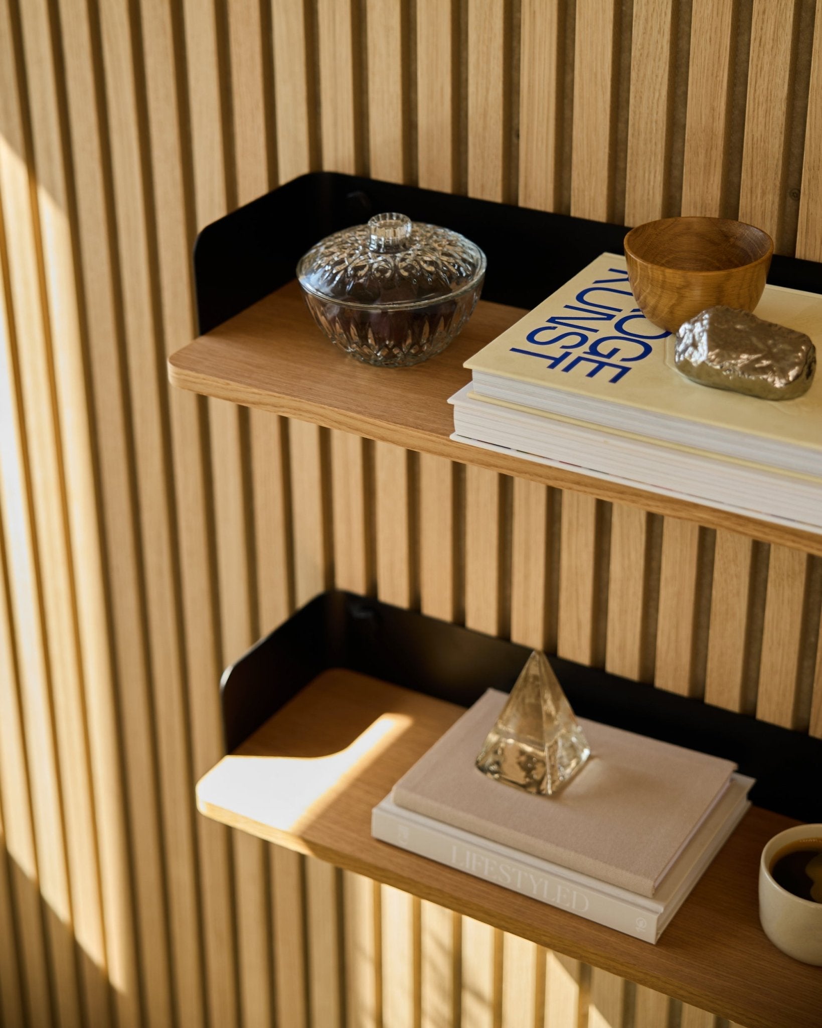 A estante Linn Bookshelf da WoodUpp em Natural Oak (55,9 cm) acomoda um frasco de vidro, uma chávena, uma pirâmide dourada e livros, sobre painéis acústicos de madeira Akupanel à luz do sol.