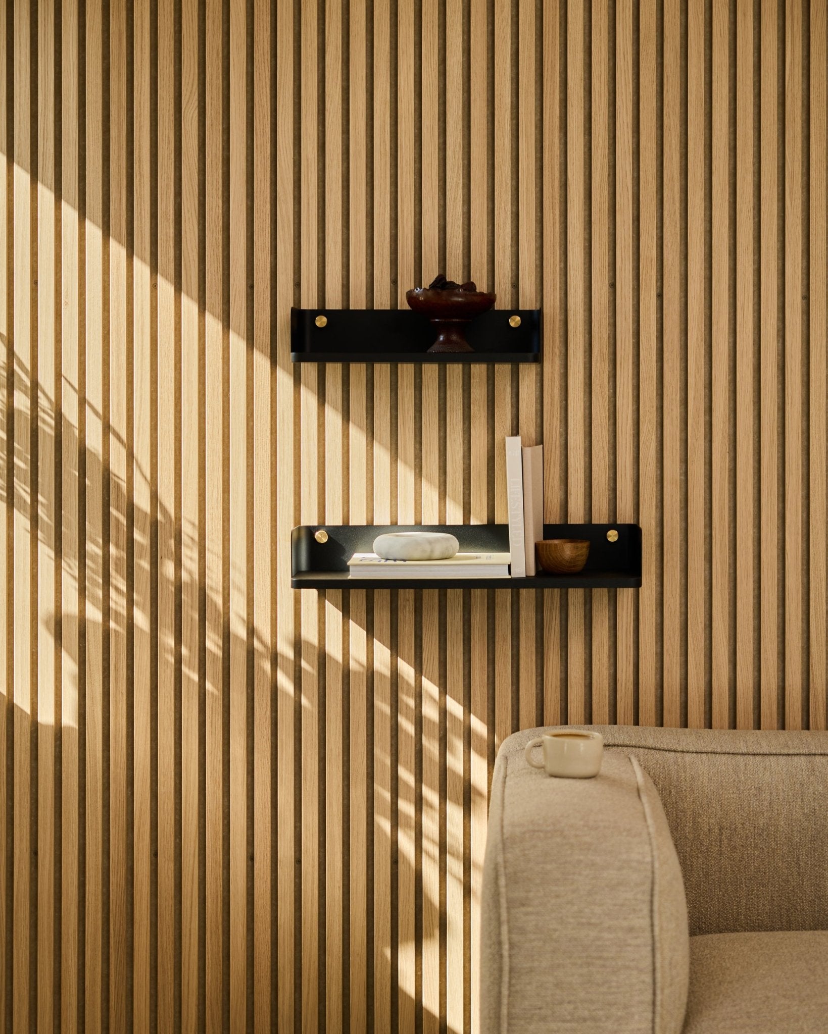 A luz do sol ilumina um sofá beige e a estante Linn da WoodUpp em black oak, 55,9 cm, colocados diante de um painel ripado nórdico minimalista Painel acústico.