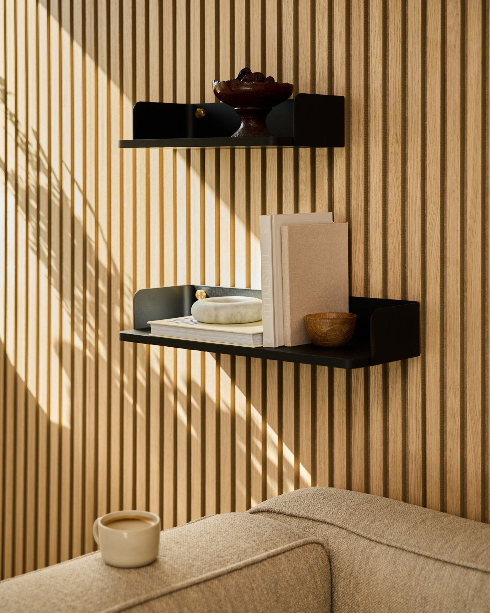 Uma estante WoodUpp black oak (55,9 cm) exibe livros, objetos decorativos e uma taça sobre uma parede com painéis acústicos de madeira; um sofá beige e uma caneca surgem em primeiro plano.