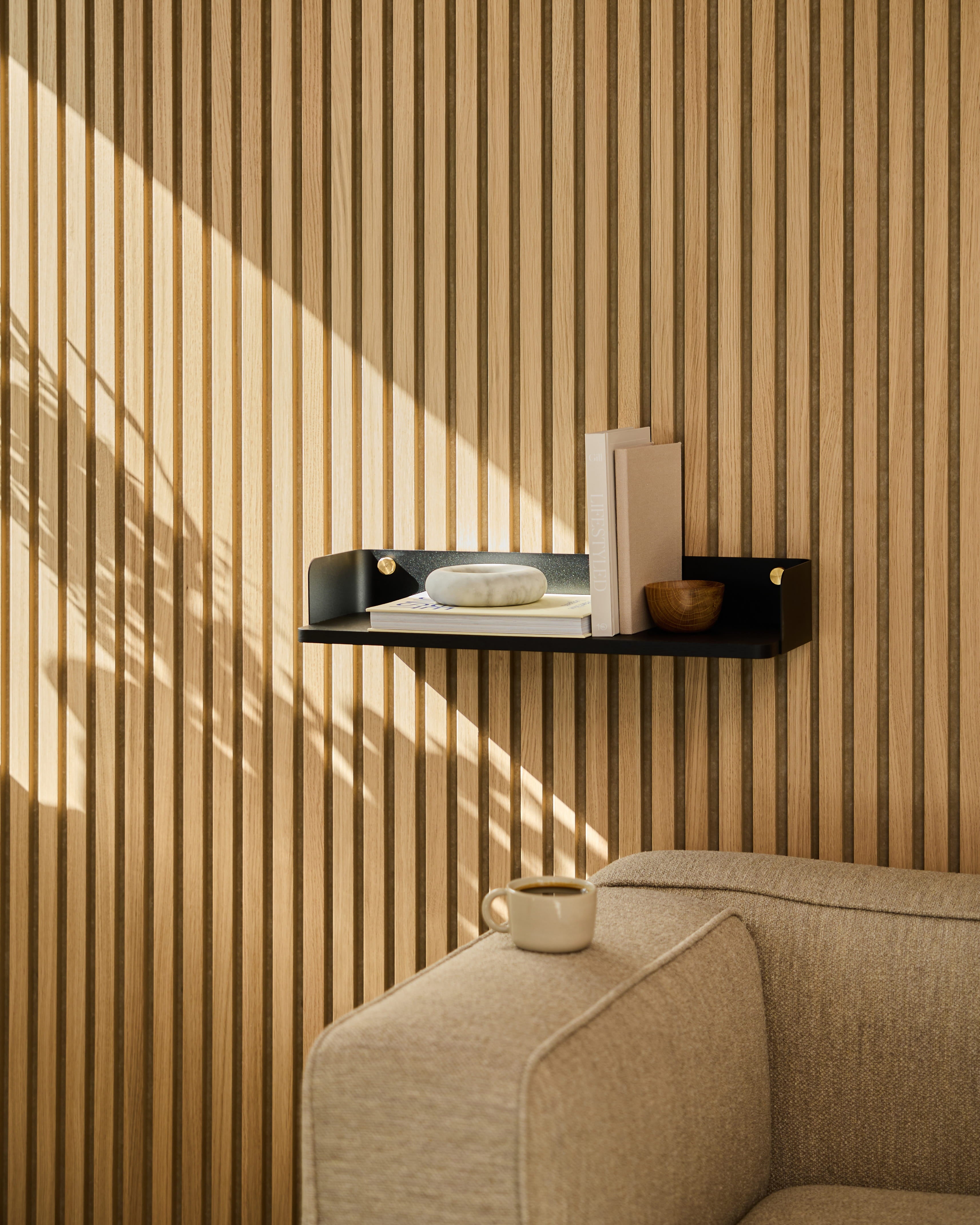 Uma estante Linn | Bookshelf / Black Oak da WoodUpp exibe objetos decorativos por cima de um sofá bege. A luz do sol projeta sombras na parede revestida com painéis acústicos de madeira às riscas.