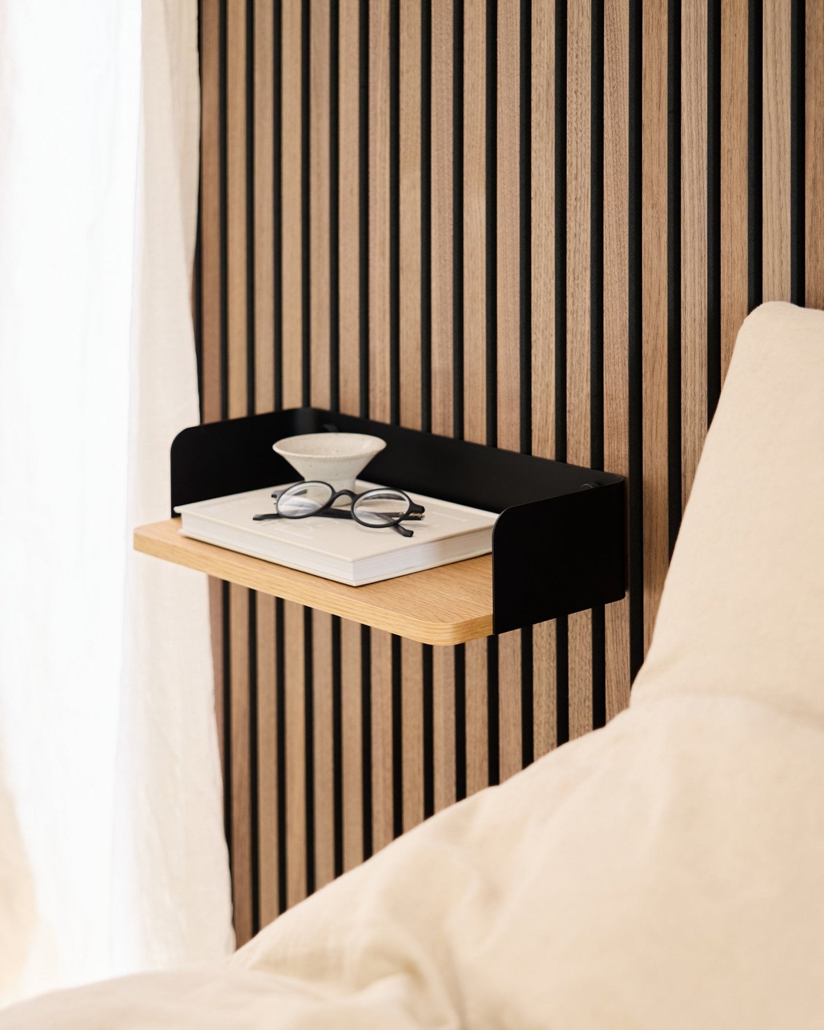 A estante Linn da WoodUpp em natural oak (39,9 cm), com um livro, óculos e um copo ao lado de uma cama, sobre um fundo de painel acústico de madeira em Akupanel.