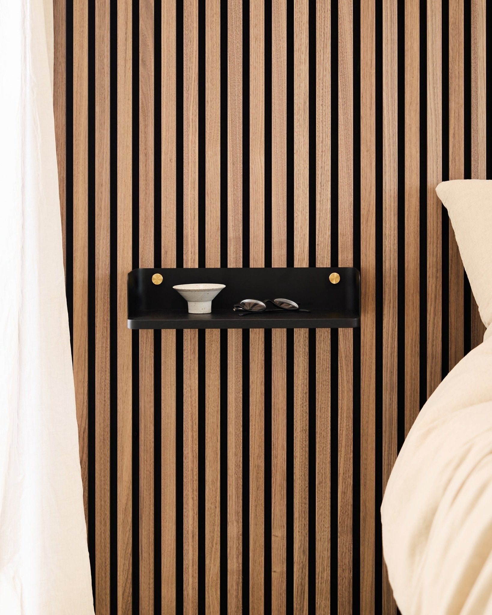 A prateleira Linn em Black Oak, da WoodUpp, está montada num painel acústico ripado de estilo nórdico, com uma taça e copos ao lado de uma cama com roupa de cama em beige.