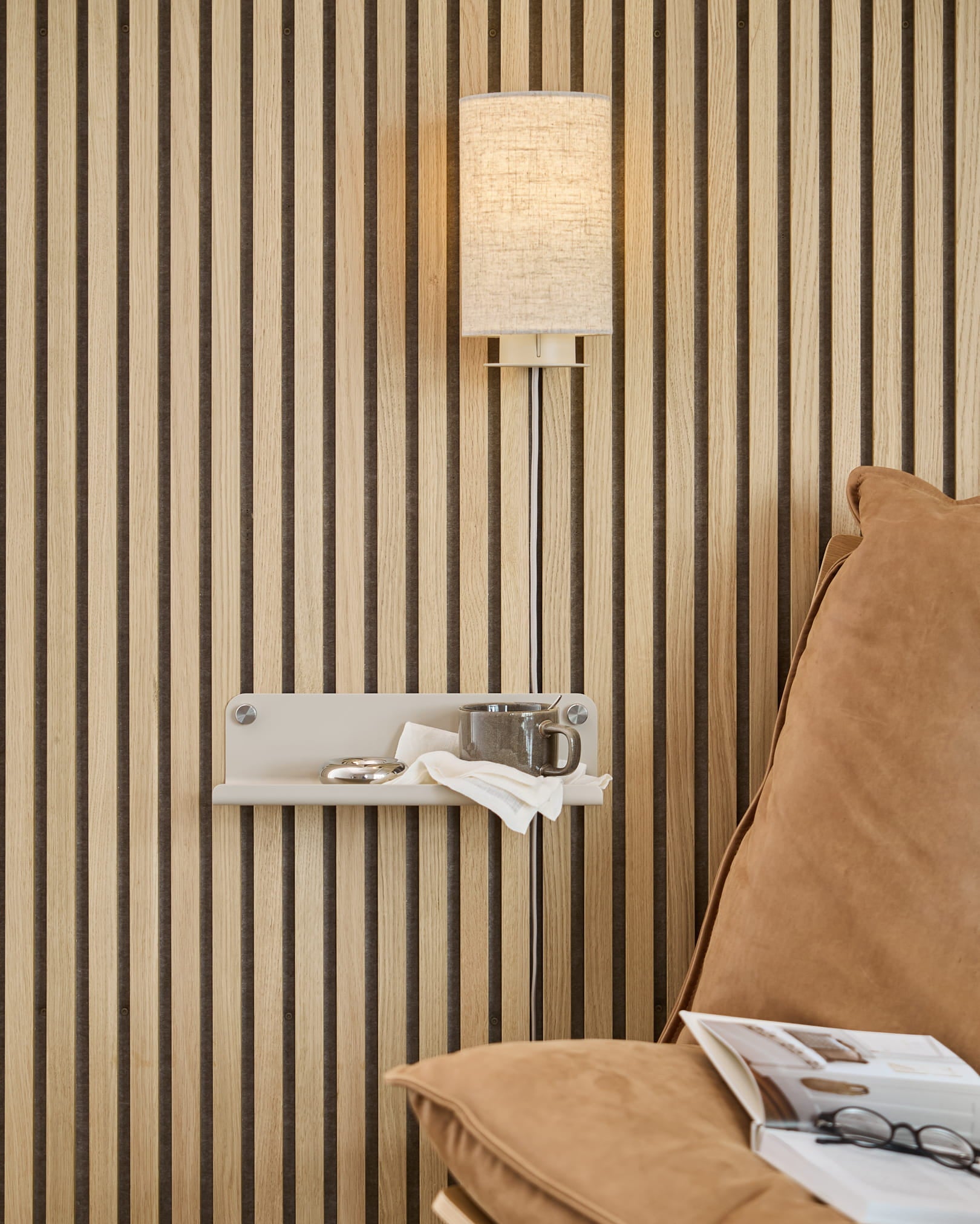 Parede com ripas acústicas verticais de madeira Akupanel, luminária de parede bege, pequena prateleira com xícara e guardanapo, sofá bege-claro com revista aberta.