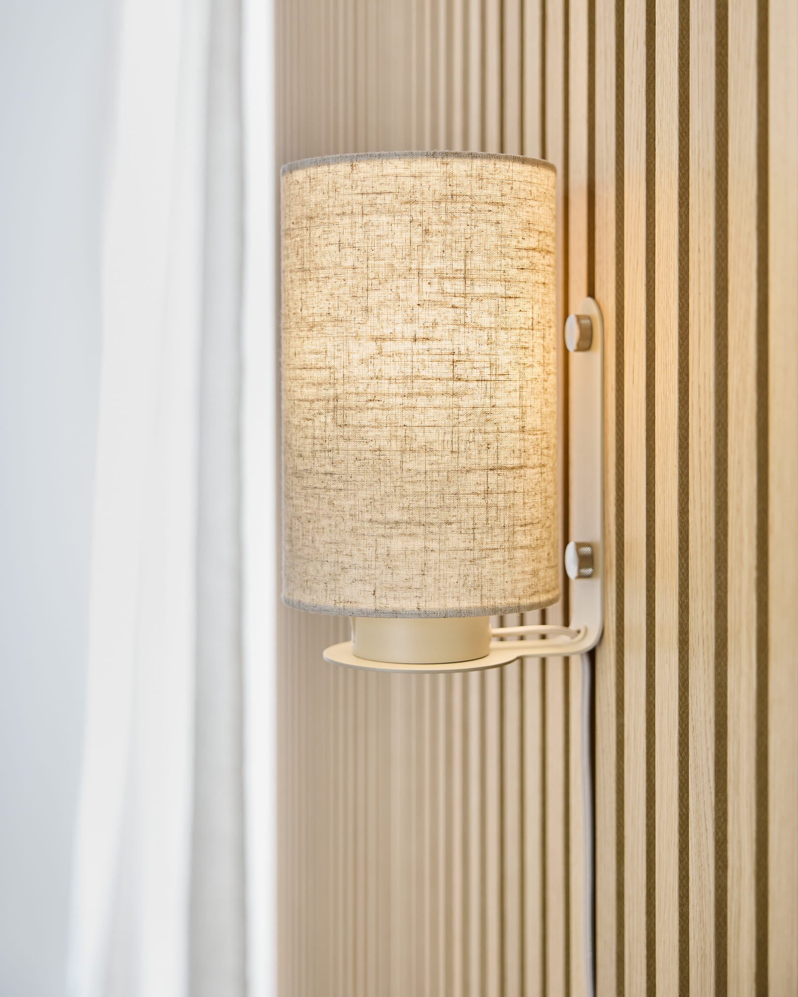 O candeeiro de parede Feya em Beige da WoodUpp projeta uma luz quente sobre uma parede acústica ripada beige Akupanel, colocada junto a uma cortina branca e a um abajur em linho-algodão.