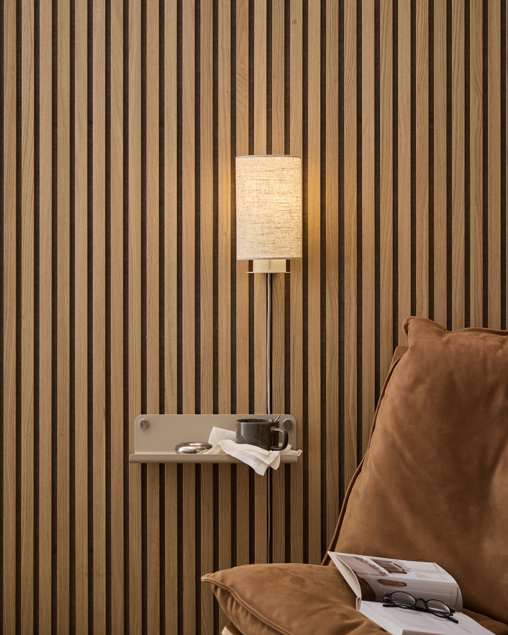 Um candeeiro de parede Feya da WoodUpp em linho-algodão beige, montado acima de um cadeirão acolhedor e de uma mesa de apoio com livro e chávena, sobre um fundo nórdico de ripas de madeira Painel acústico.