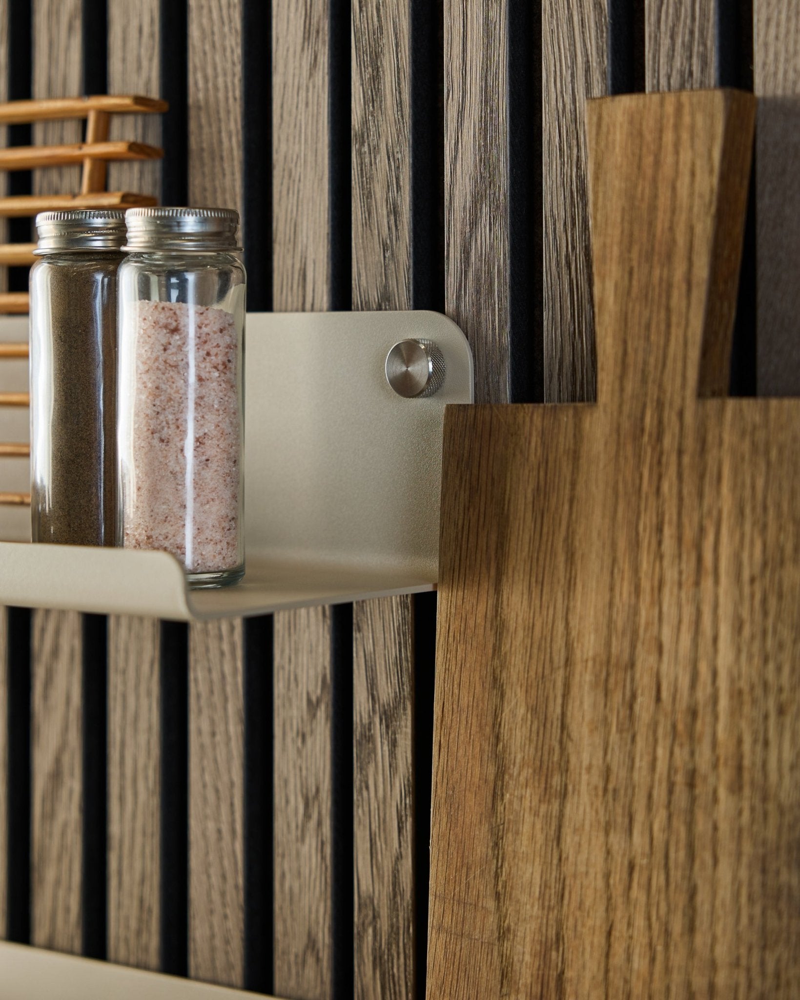 Frascos de vidro com especiarias e uma tábua de cortar em madeira pousam na prateleira larga WoodUpp Norr em beige, 35,9 cm, montada sobre um painel acústico de ripas vertical.