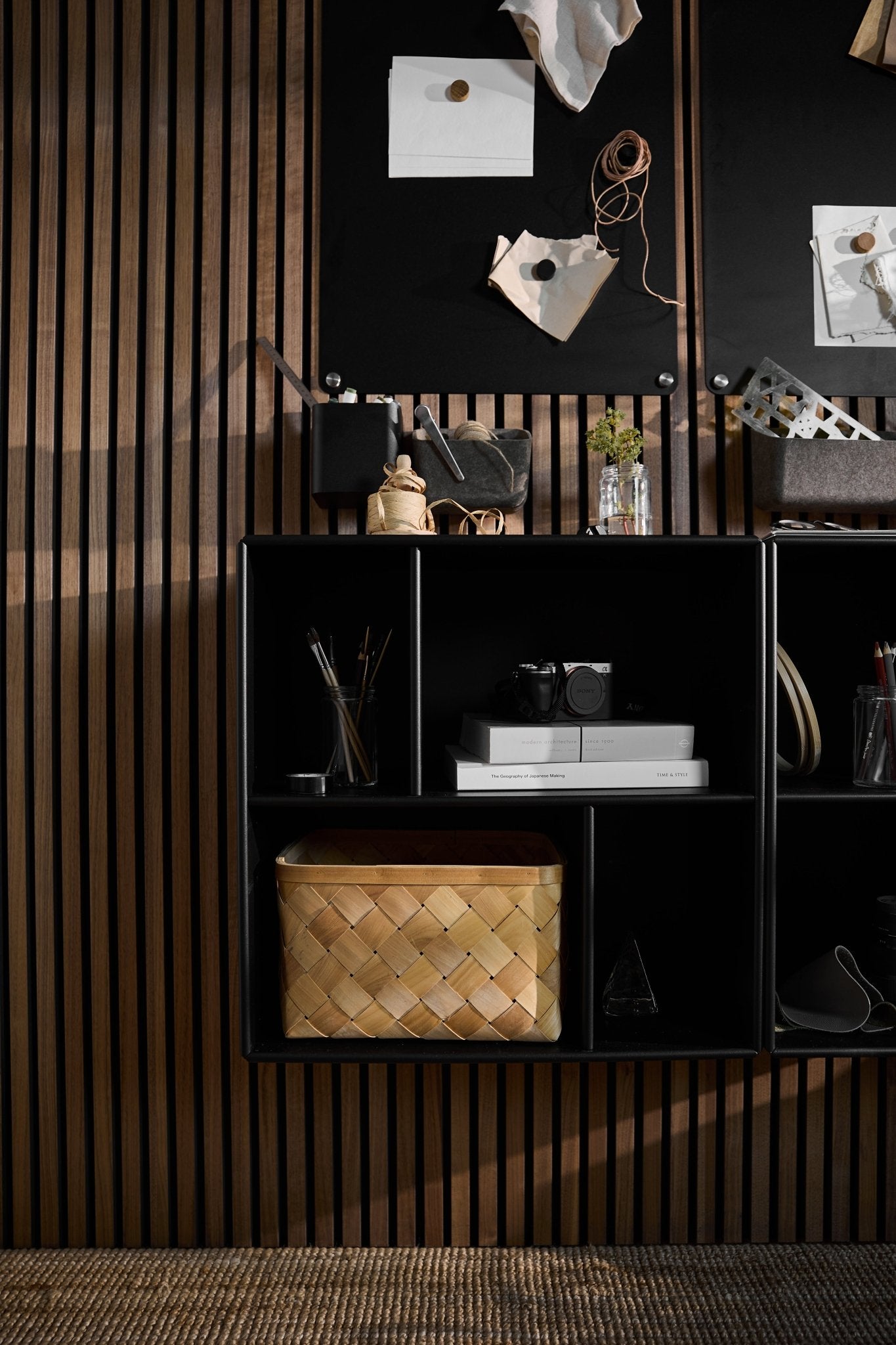 O armário aberto Arka em preto da WoodUpp, situado por baixo de um quadro de avisos preto, livros, uma câmara, objetos decorativos e um cesto de vime, tendo como pano de fundo um painel acústico nórdico com ripas.