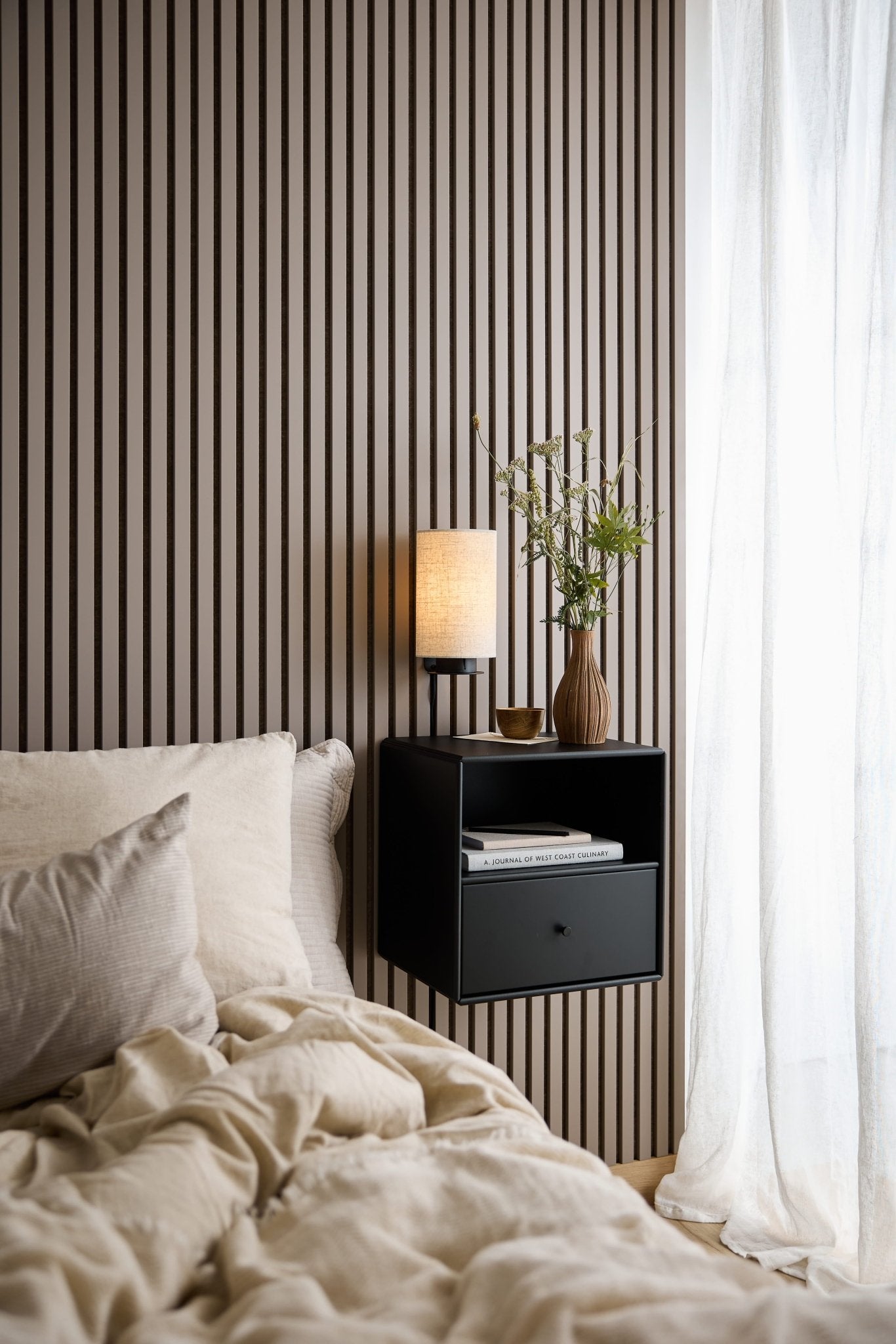 Cama com roupa de cama bege, gaveta da estante «WoodUpp Arka» preta, candeeiro e vaso junto a uma janela com cortinas brancas transparentes.