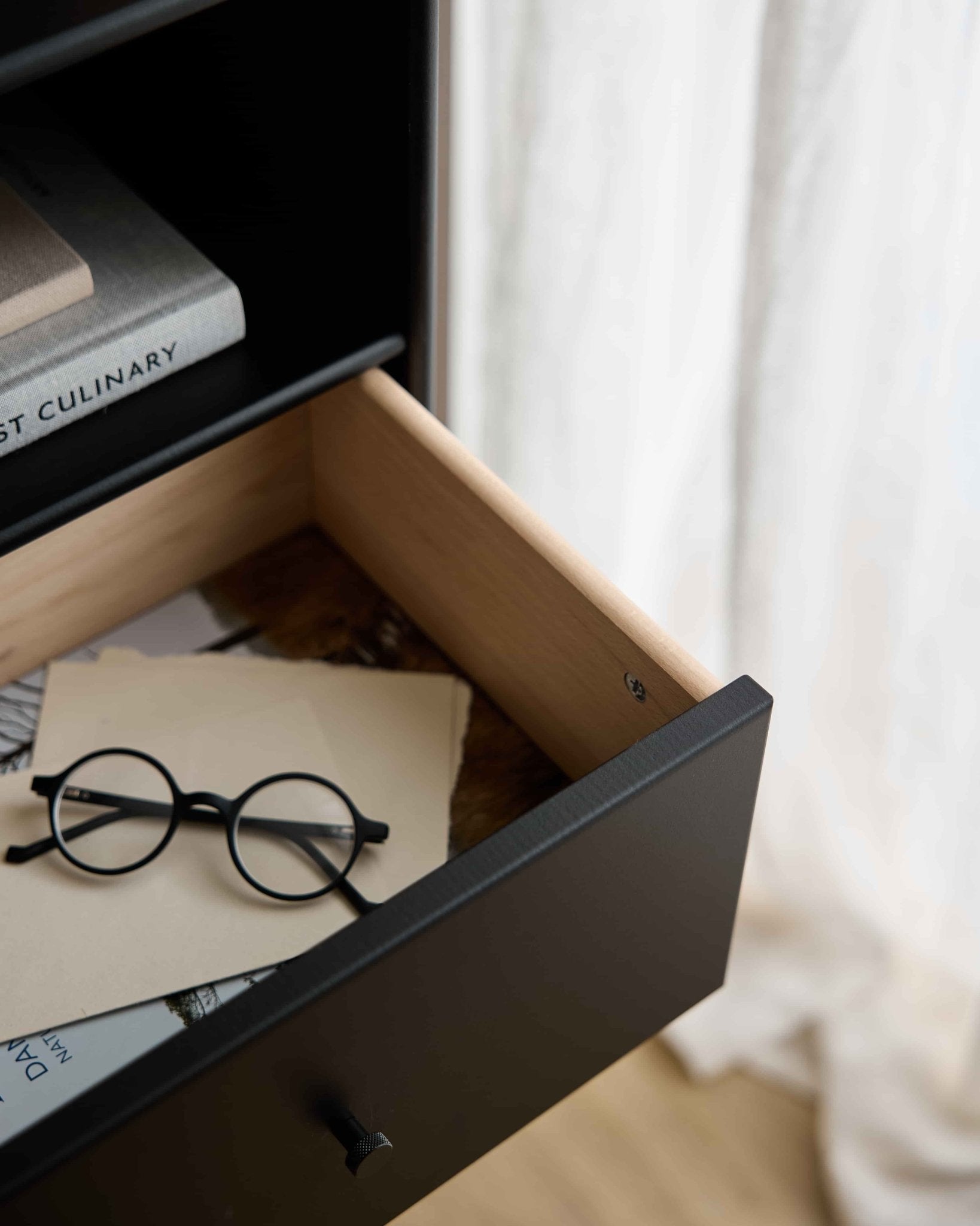 Óculos redondos pretos e papéis numa gaveta aberta da estante WoodUpp Arka | Gaveta para estante / Preta, por baixo dos livros, com cortinas transparentes a difundir suavemente a luz de fundo.