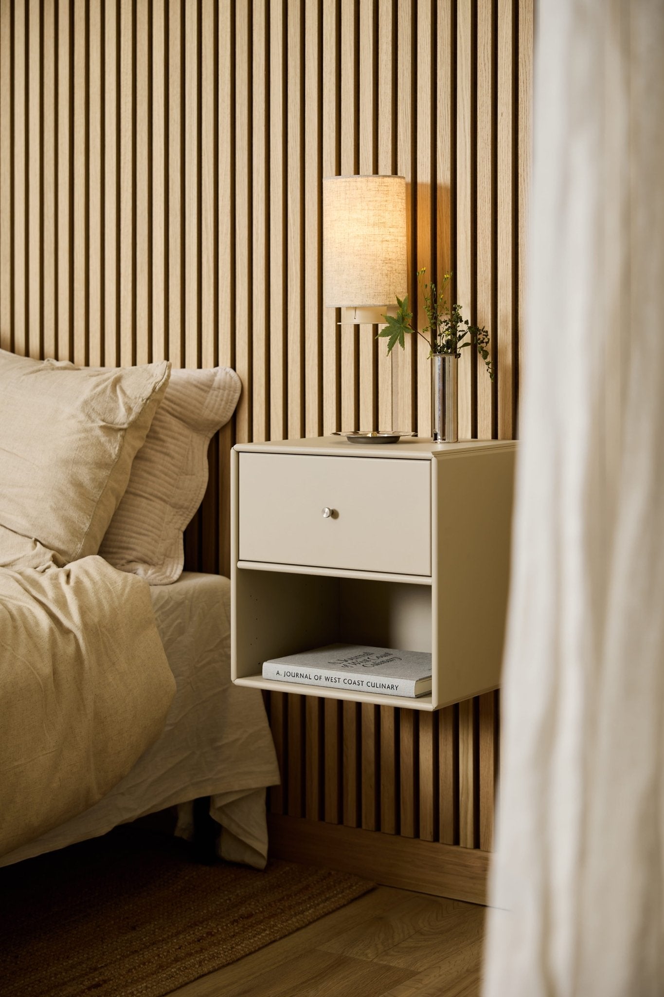 Uma estante com gaveta WoodUpp Arka, na cor bege, junto à cama, tem um candeeiro, livros e um vaso, tendo como pano de fundo um painel acústico de ripas de madeira WoodUpp Akupanel.