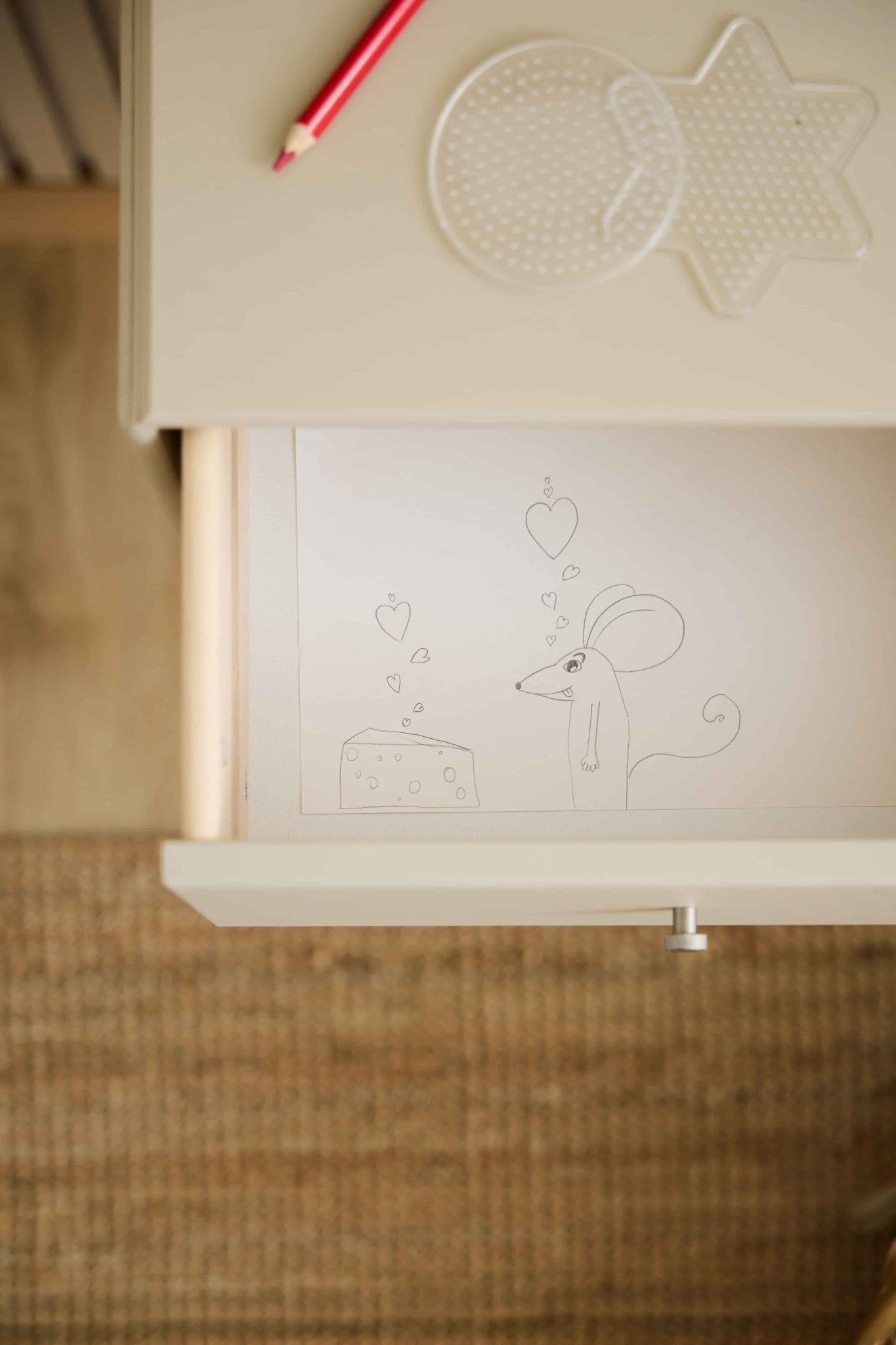 Um rato e um queijo com motivos em forma de coração decoram uma gaveta da estante WoodUpp Arka em bege; um lápis vermelho e painéis perfurados repousam sobre o tampo da mesa a condizer, acima.