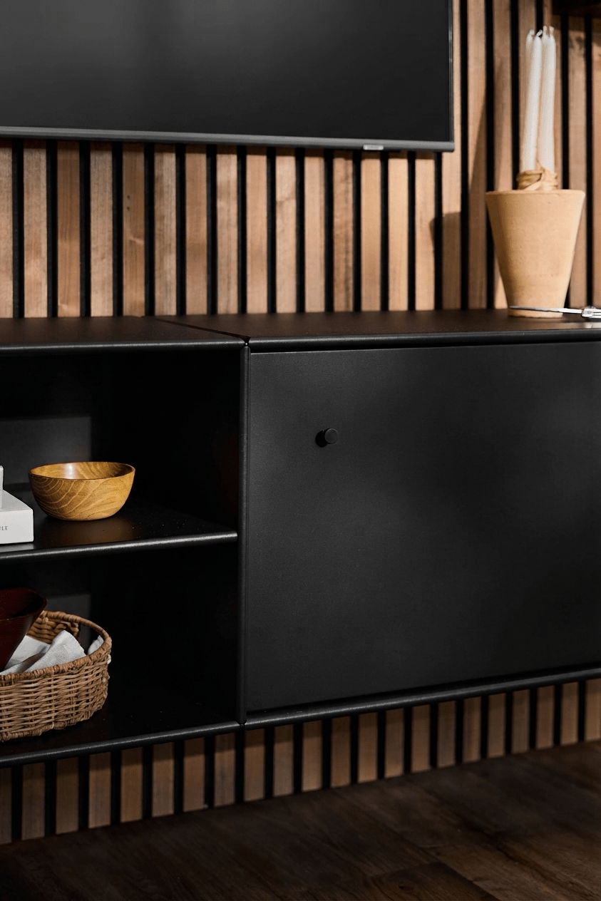 Móvel de televisão «WoodUpp Arka» em preto, com prateleiras abertas para livros e cestos, vaso em tom castanho-claro, sistema inteligente de organização de cabos e painel acústico vertical em madeira «Akupanel» como pano de fundo.