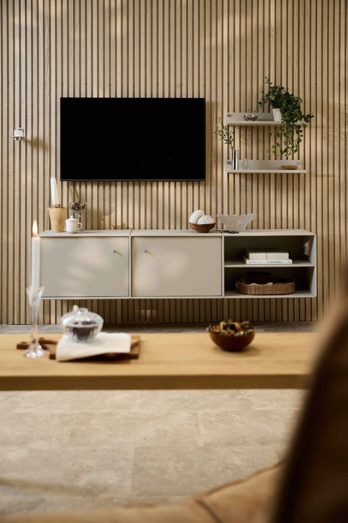 Sala de estar moderna com o móvel de televisão «Arka» da WoodUpp, conjunto de assentos modulares, prateleiras de estilo nórdico com plantas e objetos decorativos, e uma vela na mesa de centro.