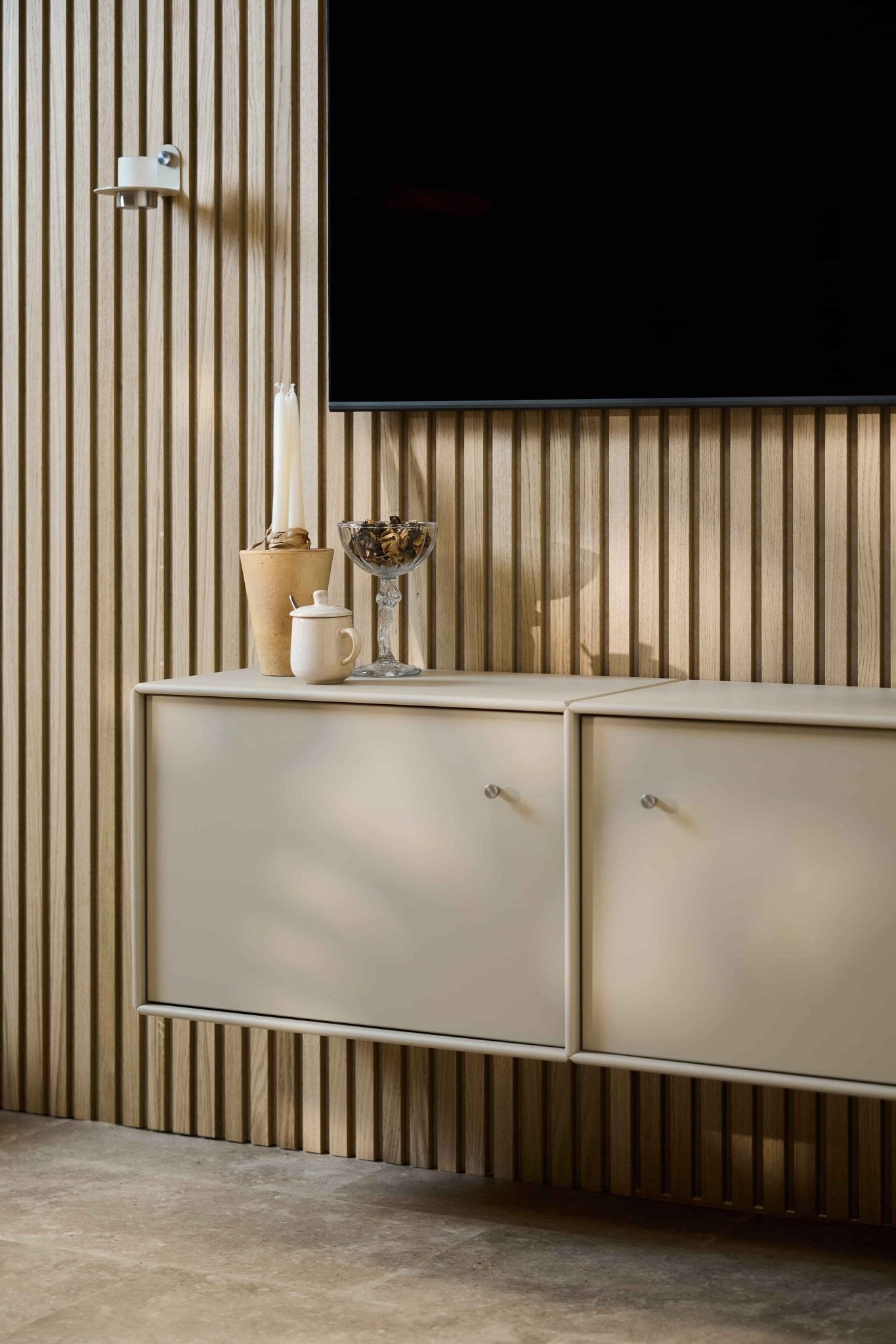 Sala de estar minimalista com a consola de TV WoodUpp Arka em bege, parede revestida com painéis acústicos de madeira Akupanel, televisão montada na parede, velas, taça de vidro, jarro branco e cabos bem organizados.