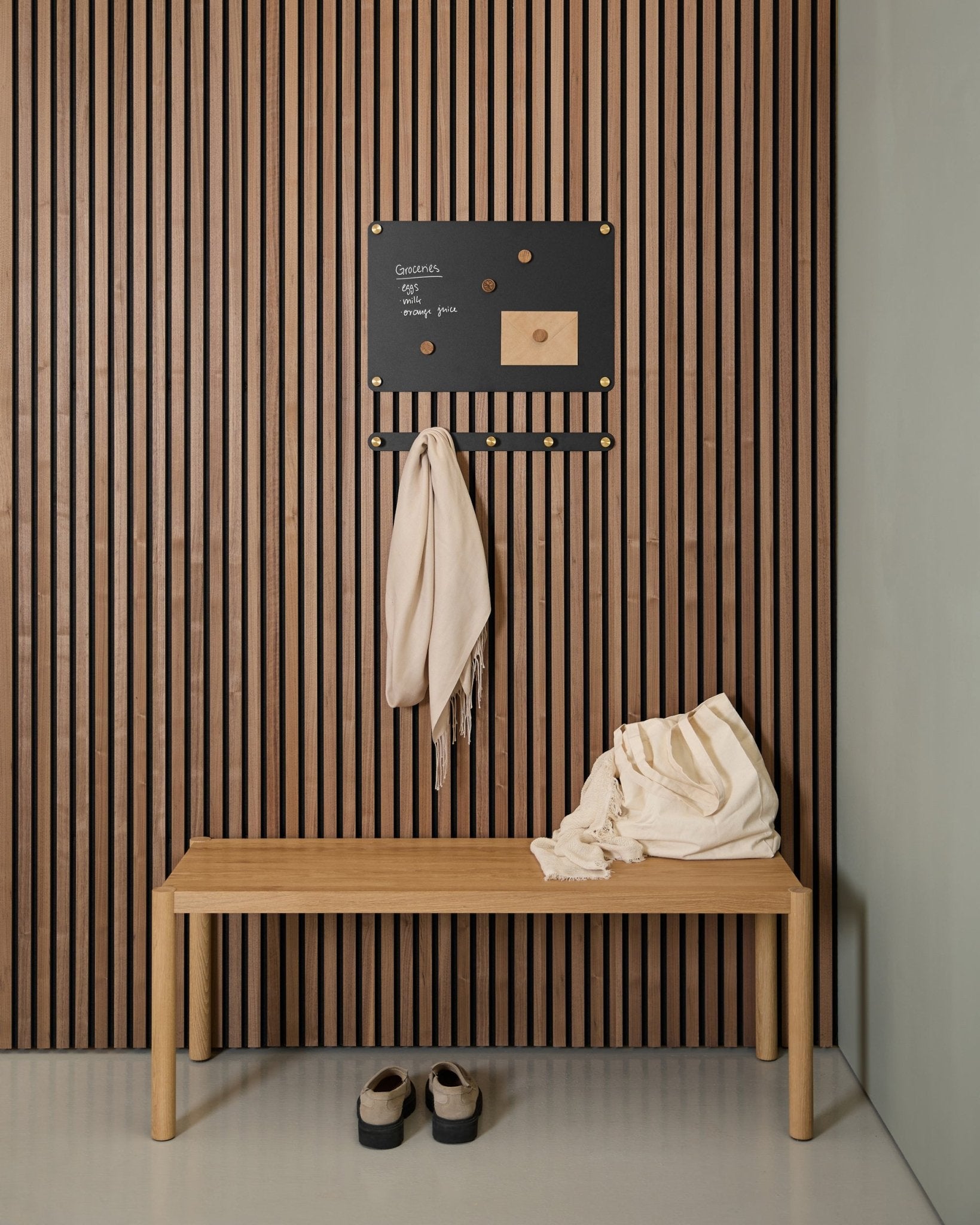 Entrada minimalista com um banco de madeira, dois cachecóis, sandálias e um small Quadro Magnético WoodUpp Norr num painel acústico ripado de madeira Akupanel.