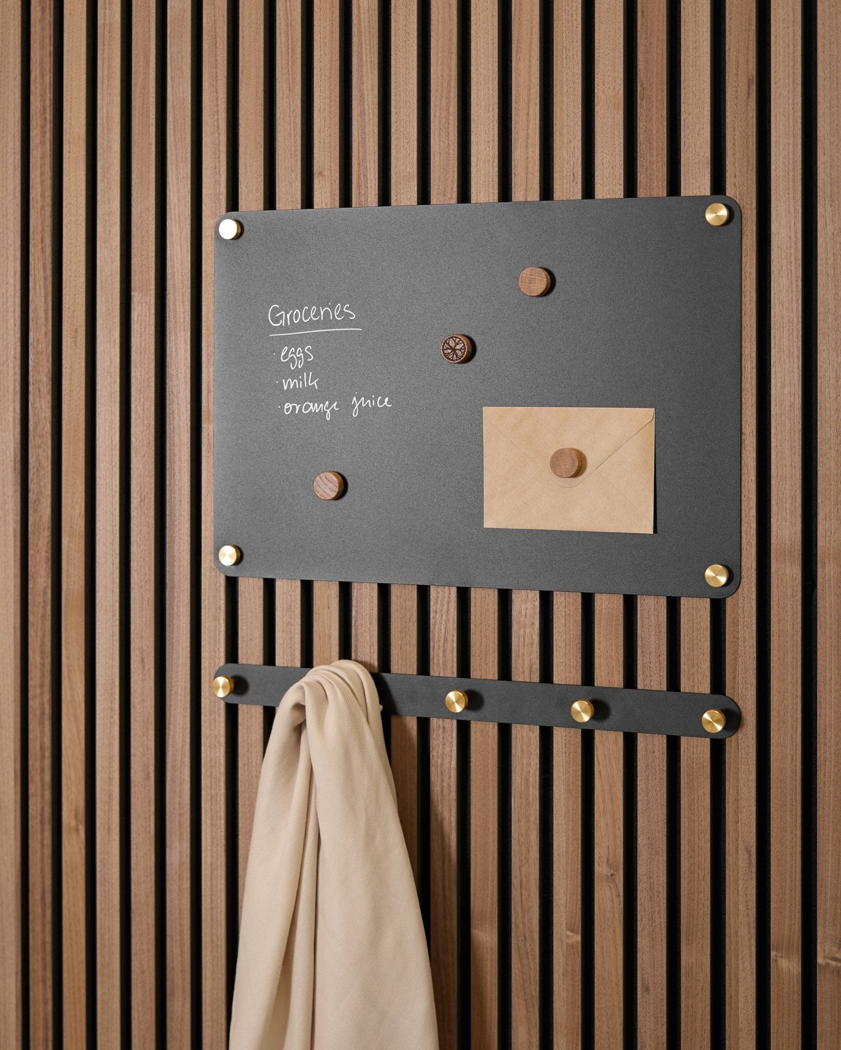 Um small black Norr Quadro Magnético da WoodUpp segura uma lista de compras, um envelope, ímanes e um beige pano em ganchos, montado num Akupanel Painel acústico.