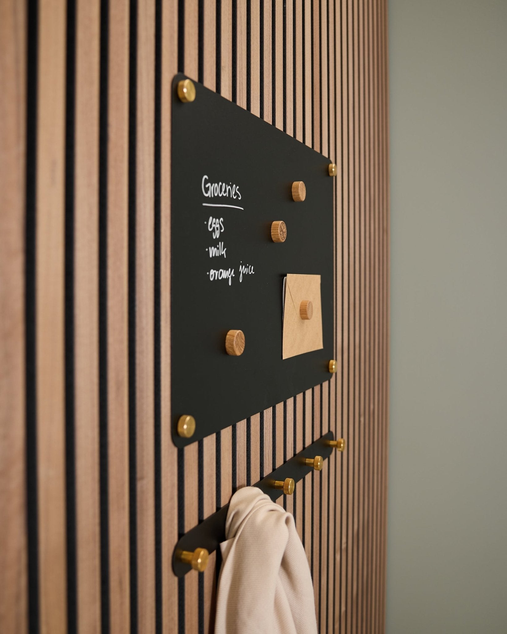 Um small black Norr Quadro Magnético da WoodUpp montado num painel acústico de madeira Akupanel exibe uma lista de compras e uma nota, com ganchos por baixo a segurarem um pano claro.