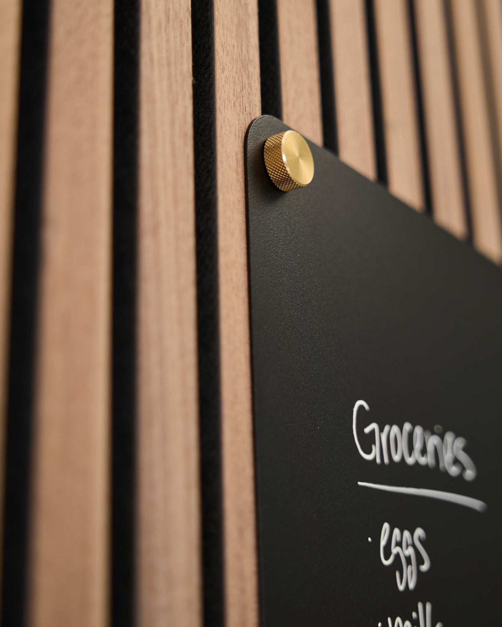 Um quadro black com uma lista de compras escrita à mão está montado num painel acústico ripado WoodUpp com dois parafusos de aperto manual brass M5x10 mm.