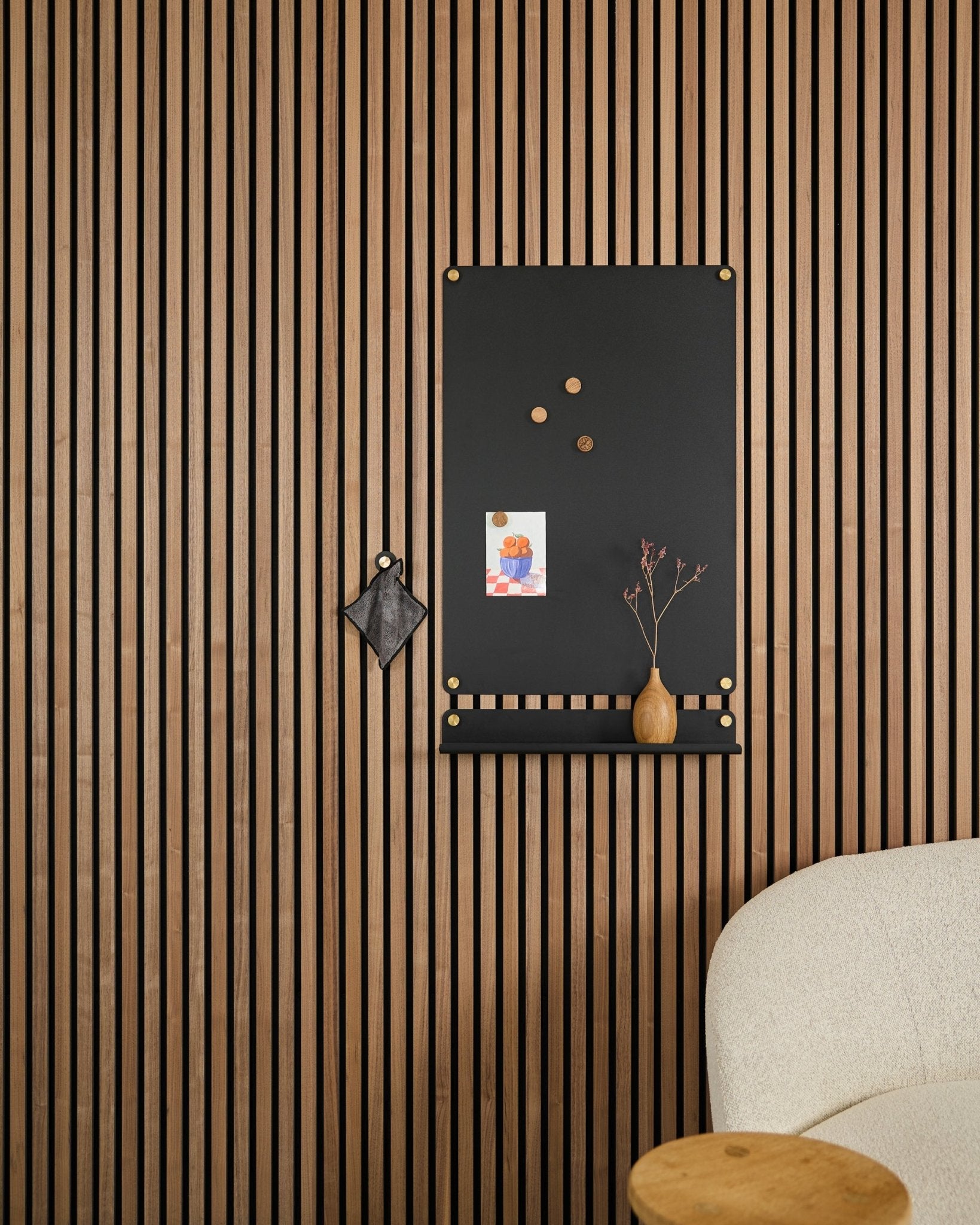 Um large black Norr Quadro Magnético da WoodUpp, concebido para painéis acústicos, está suspenso ao lado de uma cadeira estofada branca, com notas, uma jarra e um pequeno objeto decorativo.
