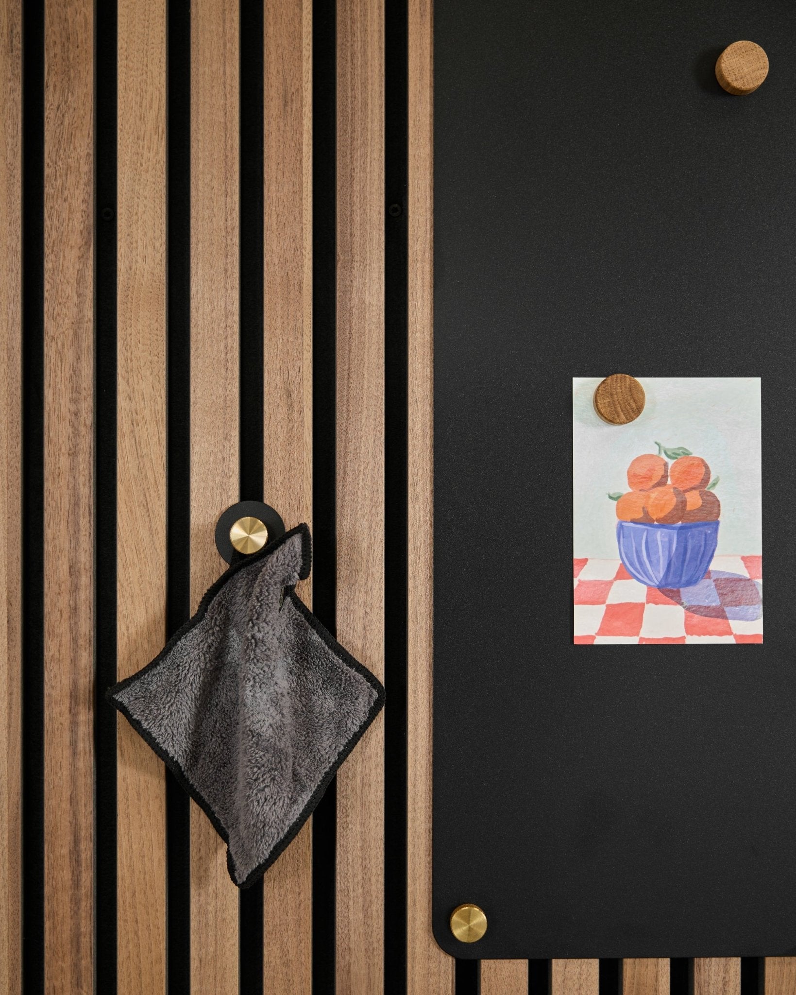 Uma toalha small está pendurada num painel acústico ripado nórdico, ao lado de um quadro black com um desenho de laranjas numa taça, preso com dois ímanes dourados.