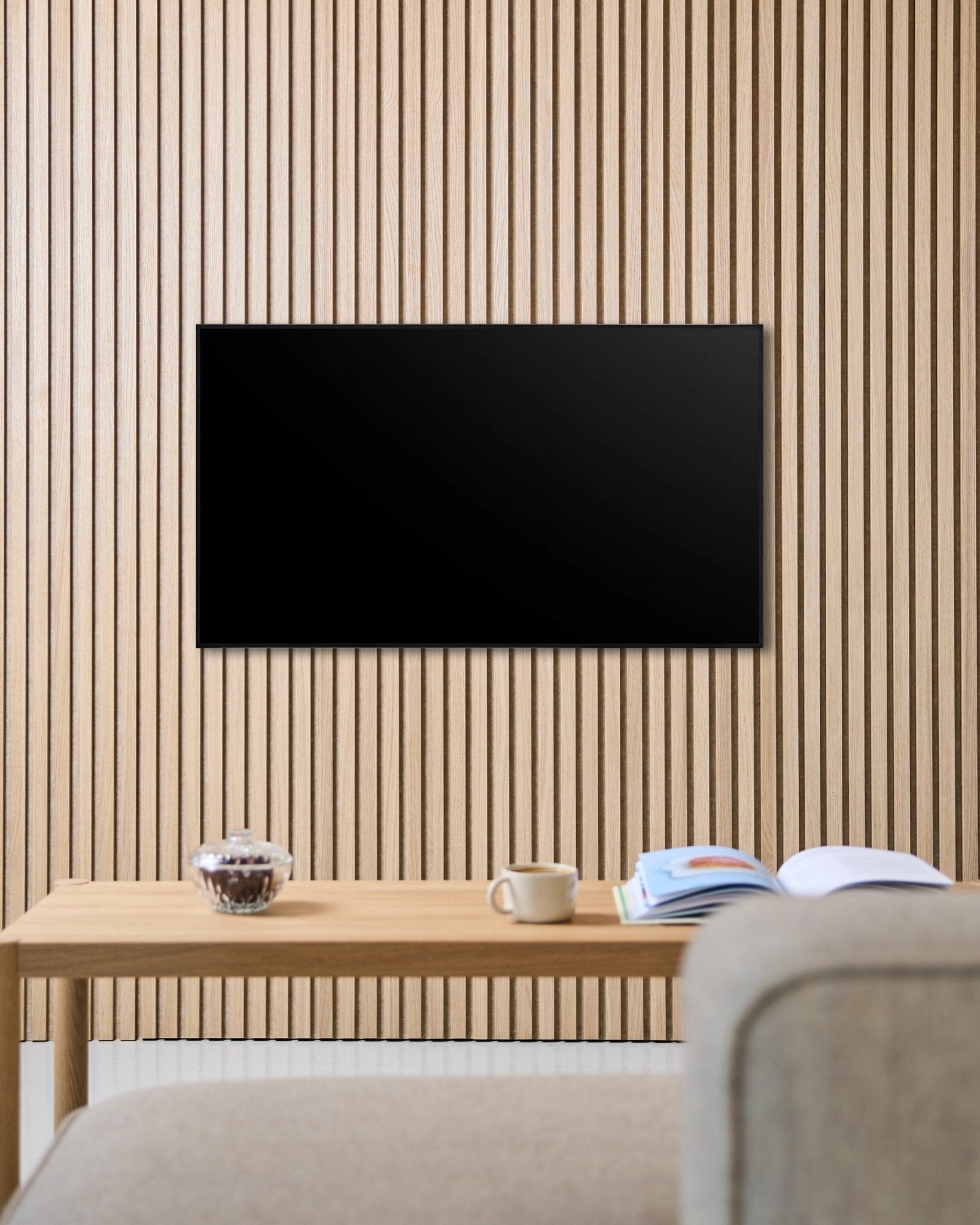 Um suporte de TV Fjell (small) da WoodUpp fixa um ecrã plano a uma parede acústica nórdica de ripas de madeira Akupanel, por cima de uma mesa com chávena, taça e revista aberta, ao lado de um sofá beige.