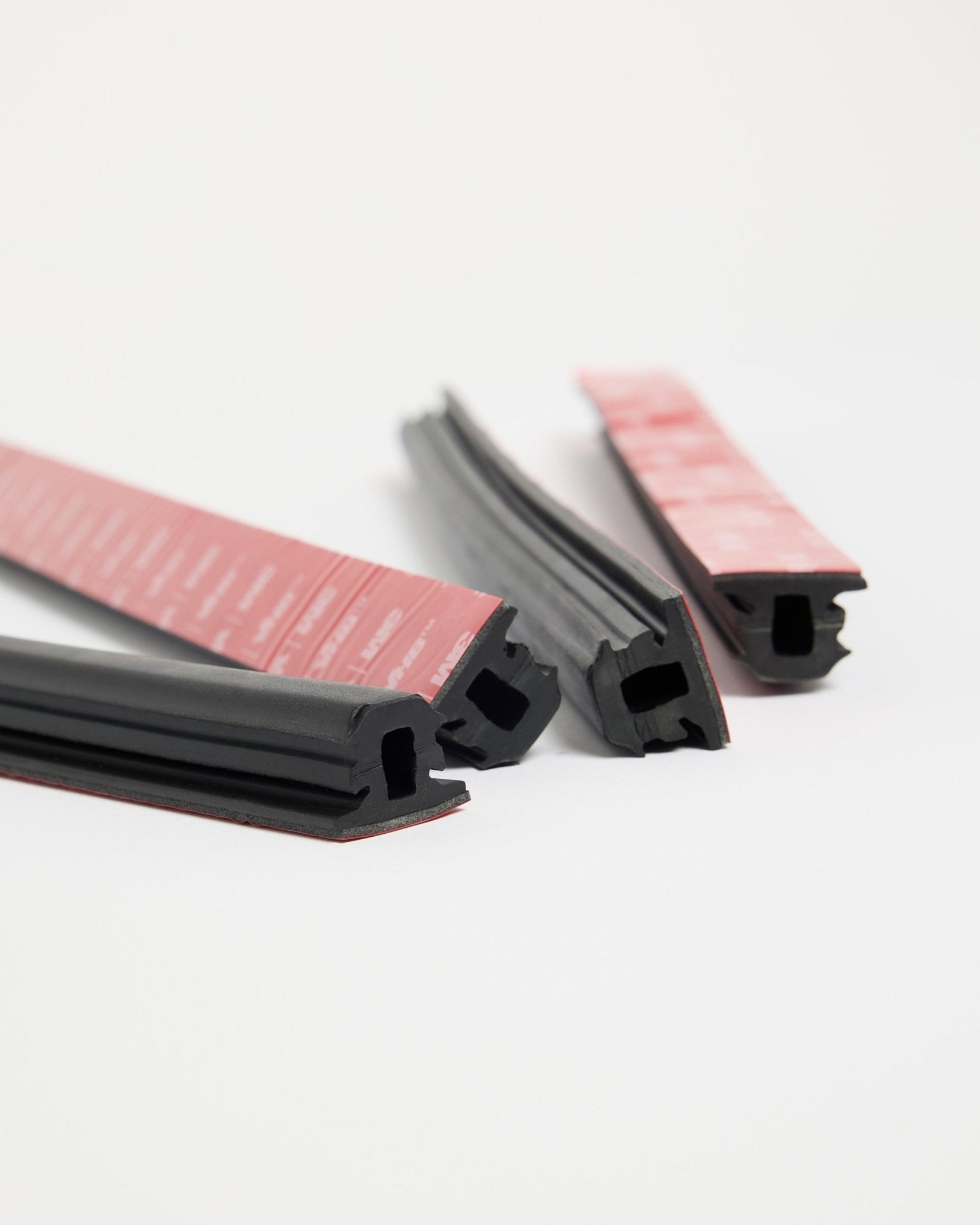 Quatro tiras de borracha WoodUpp Dupps Pop-on Profile black com adesivo vermelho, concebidas para instalação Akupanel, dispostas sobre uma superfície branca em várias direções.