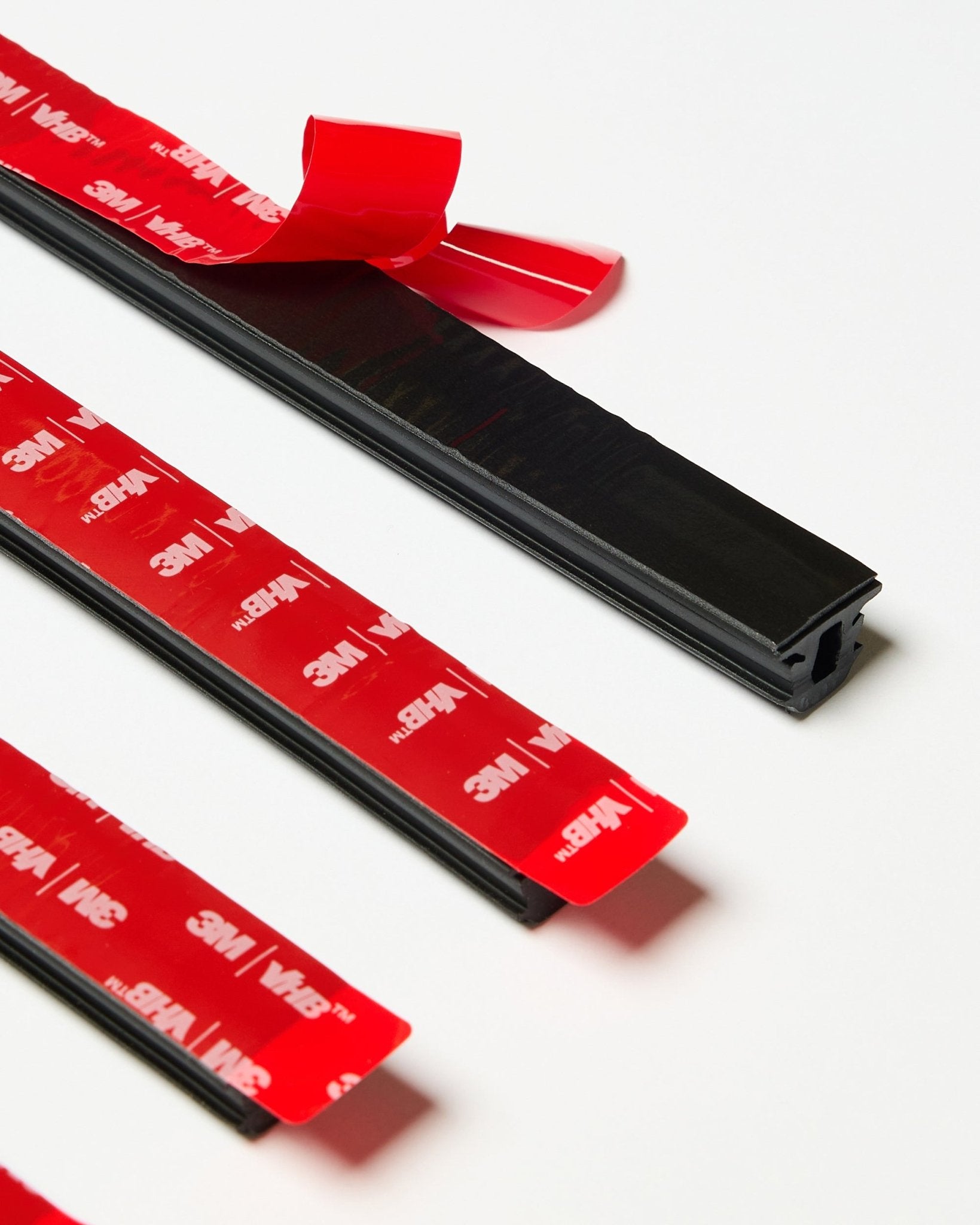 Conjunto de quatro tiras WoodUpp CREATE Pop-on Profile black com a fita adesiva vermelha 3M visível, uma tira parcialmente descolada, apresentado sobre uma superfície branca.