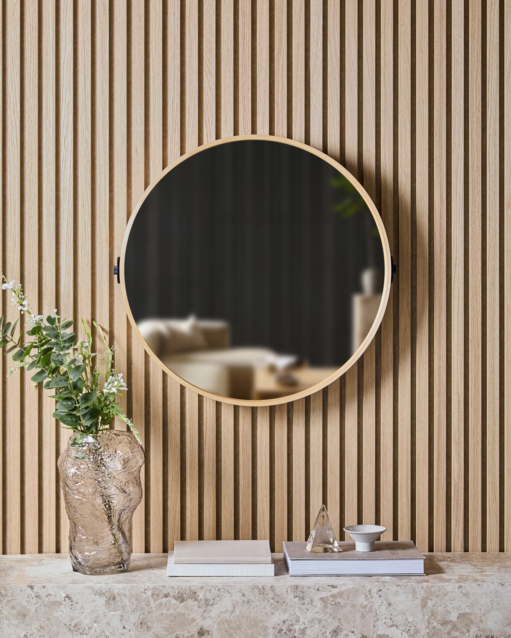 O Linn | Mirror em Natural Oak da WoodUpp está montado num painel ripado nórdico Painel acústico, por cima de uma prateleira em pedra com um vaso de vidro, livros e peças decorativas.